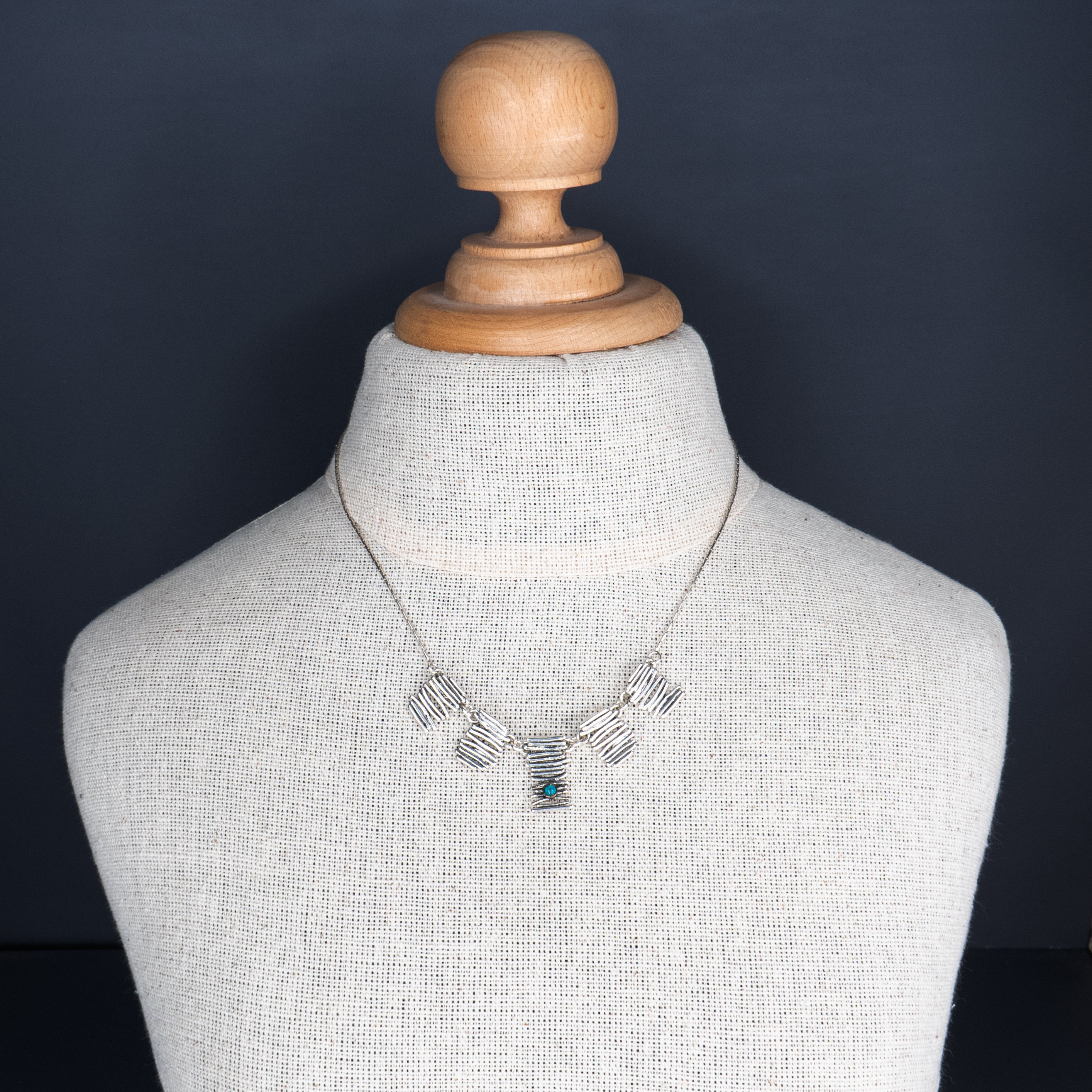 שרשרת קצרה ומרשימה עם שלושה חלקי כסף מחוספסים, בסגנון גאומטרי ואמנותי. בכל אחד מהם שובצה אבן טורקיז כחולה – סמל להגנה, שלווה וחיבור לטבע. האבן המרכזית חתוכה בצורת ריבוע, והשתיים בצדדים עגולות. הצילום ממוקד על צוואר חלק ומואר, ומדגיש את השילוב בין העיצוב המתכתי הקר לבין חמימות הצבע של הטורקיז.