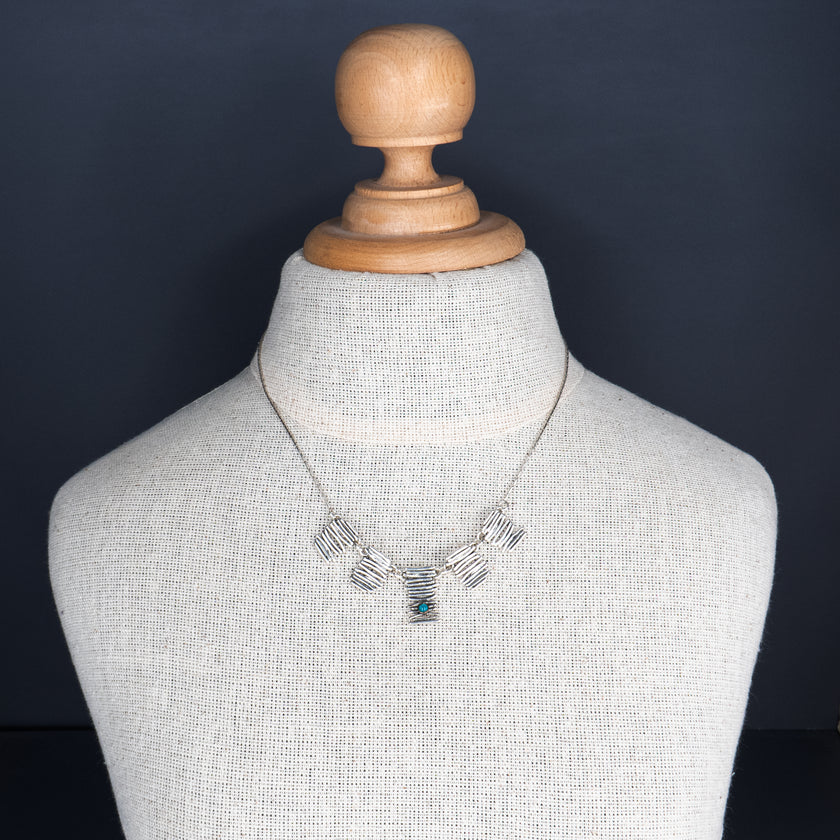 שרשרת קצרה ומרשימה עם שלושה חלקי כסף מחוספסים, בסגנון גאומטרי ואמנותי. בכל אחד מהם שובצה אבן טורקיז כחולה – סמל להגנה, שלווה וחיבור לטבע. האבן המרכזית חתוכה בצורת ריבוע, והשתיים בצדדים עגולות. הצילום ממוקד על צוואר חלק ומואר, ומדגיש את השילוב בין העיצוב המתכתי הקר לבין חמימות הצבע של הטורקיז.
