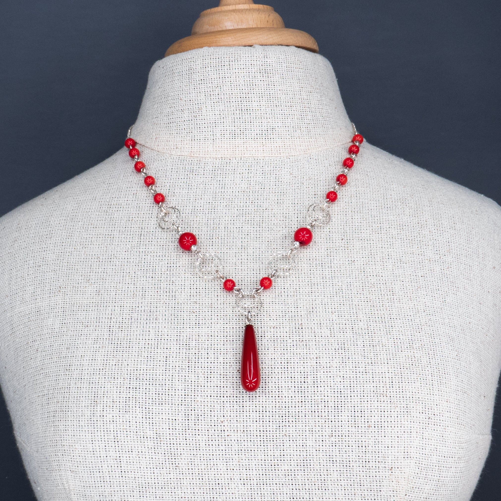 תכשיט נועז ונשי, עשוי כסף עם שילוב של חרוזים מאלמוג סינתטי בגוון אדום דובדבן. תליון טיפתי במרכז משלים את העיצוב. מוצג על צוואר חשוף בגוון עור בהיר ועם בגד ג’ינס פתוח – שילוב קונטרסטי של אלגנטיות ופשטות אורבנית.