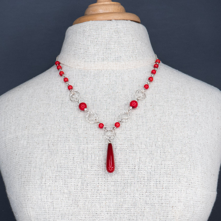 תכשיט נועז ונשי, עשוי כסף עם שילוב של חרוזים מאלמוג סינתטי בגוון אדום דובדבן. תליון טיפתי במרכז משלים את העיצוב. מוצג על צוואר חשוף בגוון עור בהיר ועם בגד ג’ינס פתוח – שילוב קונטרסטי של אלגנטיות ופשטות אורבנית.