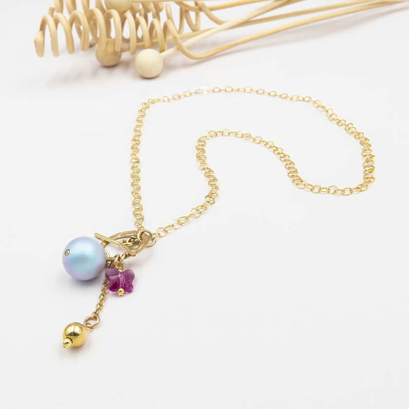 תליון ייחודי המורכב מפנינה בגוון כחול-סגלגל, חרוז פרפר ורוד ותליון מתכתי מוזהב, תלויים על סוגר טוגל מוזהב שממוקם בקדמת שרשרת חוליות זהובה. עיצוב אלגנטי עם נגיעה של פנטזיה וצבע.