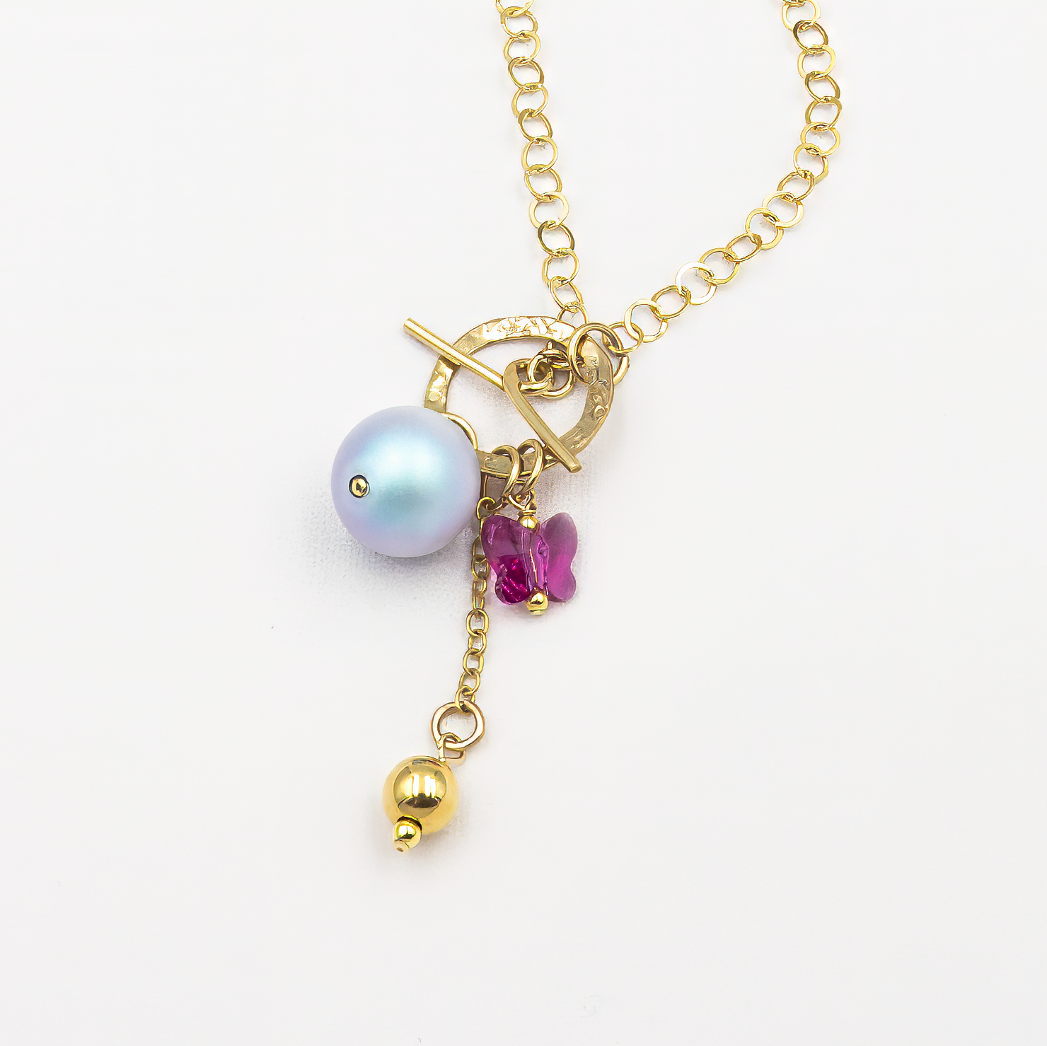 תכשיט נשי ועדין – שרשרת חוליות זהובה עם סגירת טוגל ייחודית בחלק הקדמי, שממנה תלויים שלושה אלמנטים: פנינה עגולה בצבע סגול פנינה, פרפר קטן בגוון ורוד פוקסיה ותליון זהוב כדורי. שילוב קיצי ומלא חיים.