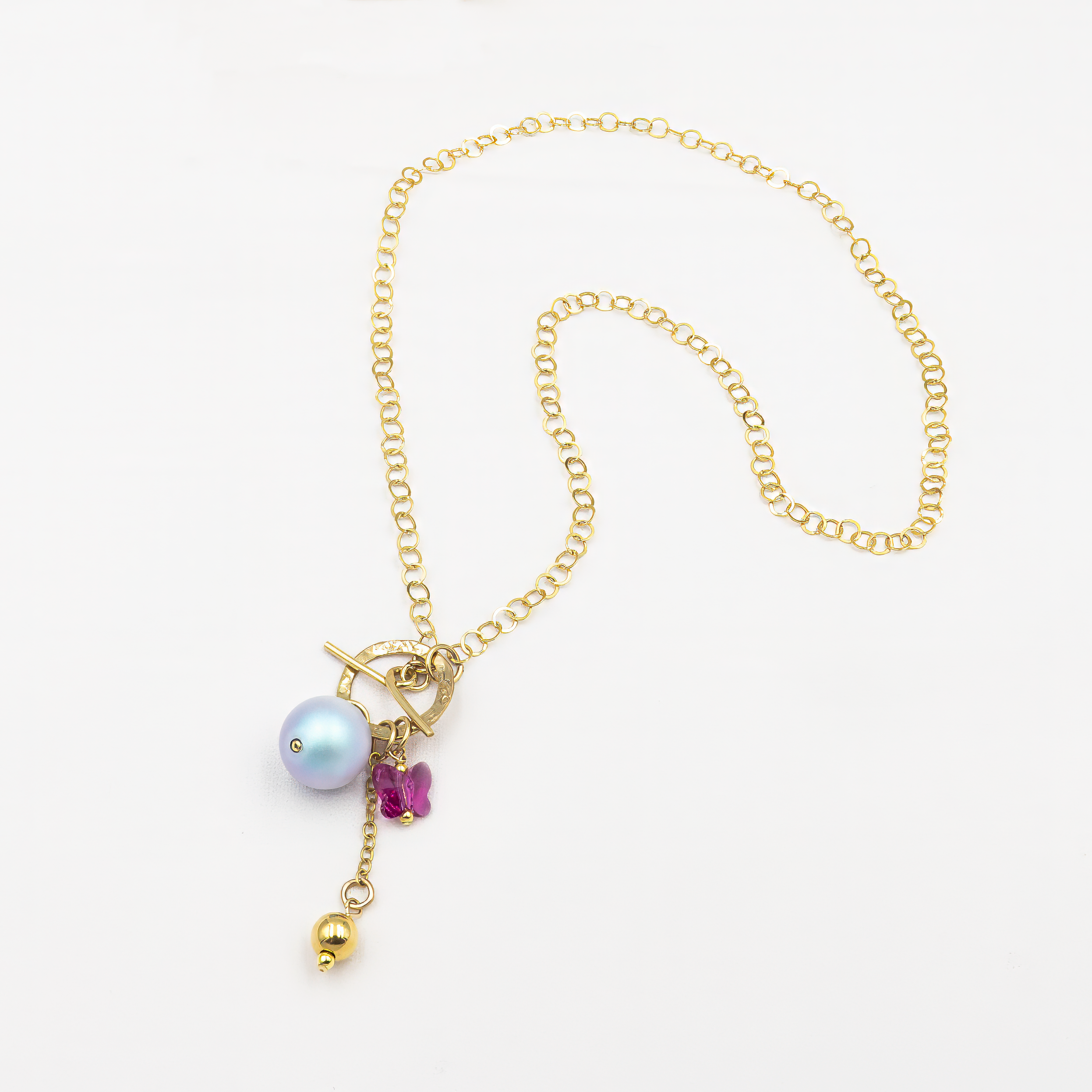 שרשרת זהב אלגנטית עם שרשרת חוליות עדינה, מעוטרת באלמנטים תלויים: פנינה בגוון סגלגל-כחול, חרוז בצורת פרפר ורוד, ותליון מתכתי קטן בגוון זהב. הסגירה הקדמית מעוצבת בסגנון טוגל, מוסיפה נוכחות אופנתית. תכשיט עם אופי שובבי אך מעודן.