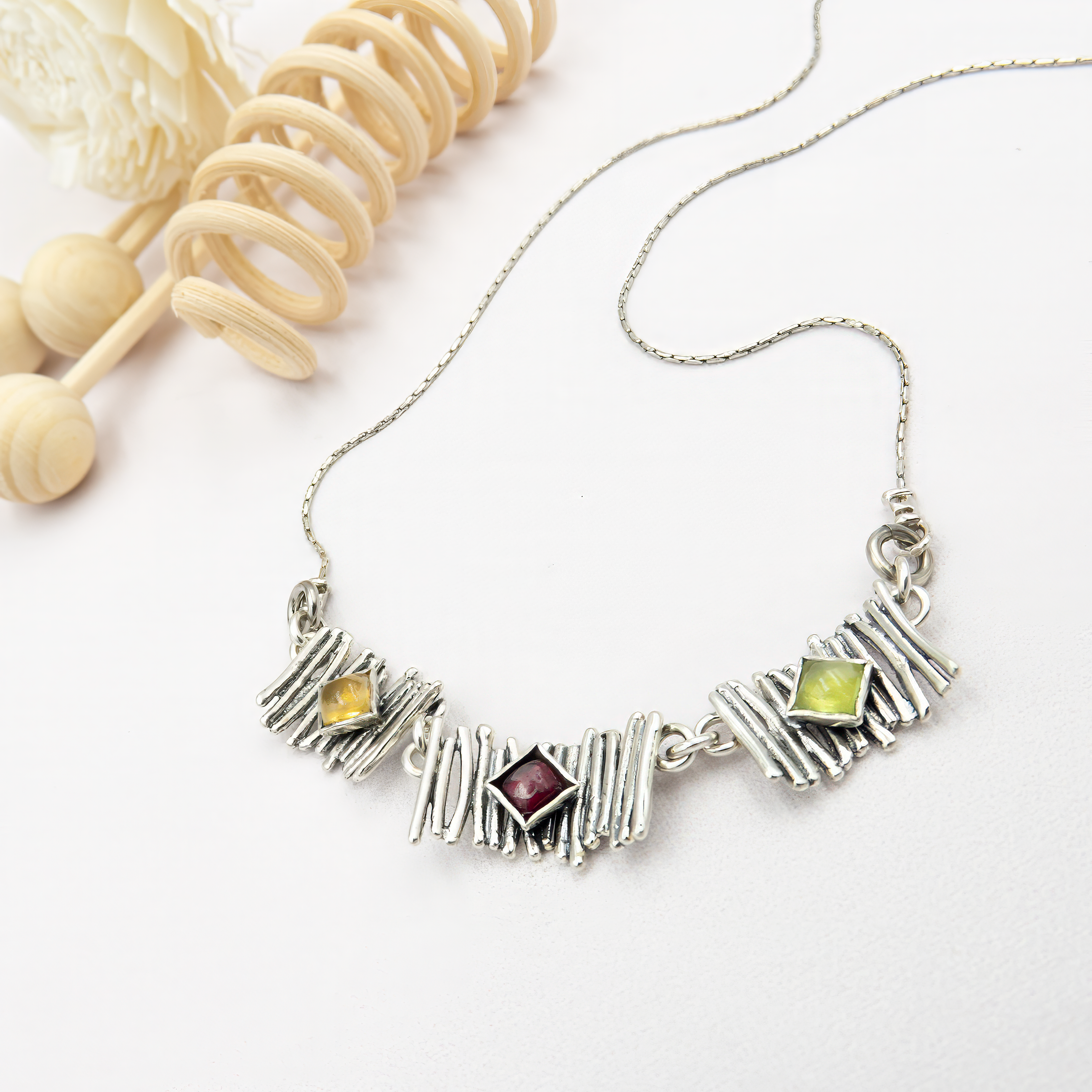 צילום תקריב של שרשרת כסף קצרה עם שלושה אלמנטים מלבניים בטקסטורה מחוספסת, המזכירה גזרי עץ. בכל אחד מהם משובצת אבן סיטרין: מרכזי בחיתוך מרובע, ושניים קטנים בצדדים בחיתוך עגול. גוון הצהוב־דבש של הסיטרינים בולט על רקע הכסף המוברש, ויוצר מראה חמים, חיובי וזוהר. השרשרת מונחת על צוואר בצבע עור ניטרלי שמבליט את הניגוד בין המתכת לאבני החן.