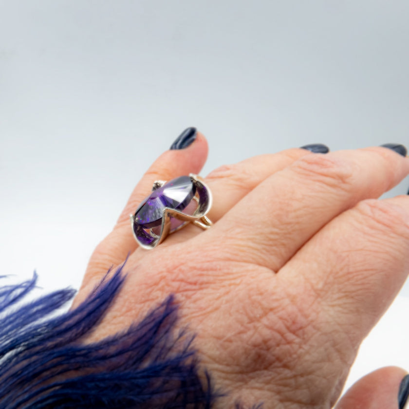 טבעת כסף בעבודת יד, כוללת בקריסטל סגול בגוון המזכיר את אבן האמטיסט – סגול מלכותי שנע בין לבנדר עדין לסגול כהה. האבן חתוכה בפאות מדויקות ויוצרת משחקי אור שמעניקים לה אור רך ומרגש. השילוב בין גוון האבן האצילי לבין מתכת הכסף המלוטשת מעניק לטבעת שילוב של רגש ואלגנטיות, בין קלאסיקה לאנרגיה מודרנית. התכשיט מתאים לנשים שמחפשות עומק, אופי רגשי עמוק שאינה מובנת מאליה. קריסטל בגוון אמטיסט מסמל איזון נפשי – ומתאים כתוספת למראה יומיומי מלא חן או כתכשיט עם משמעות לערב מיוחד.