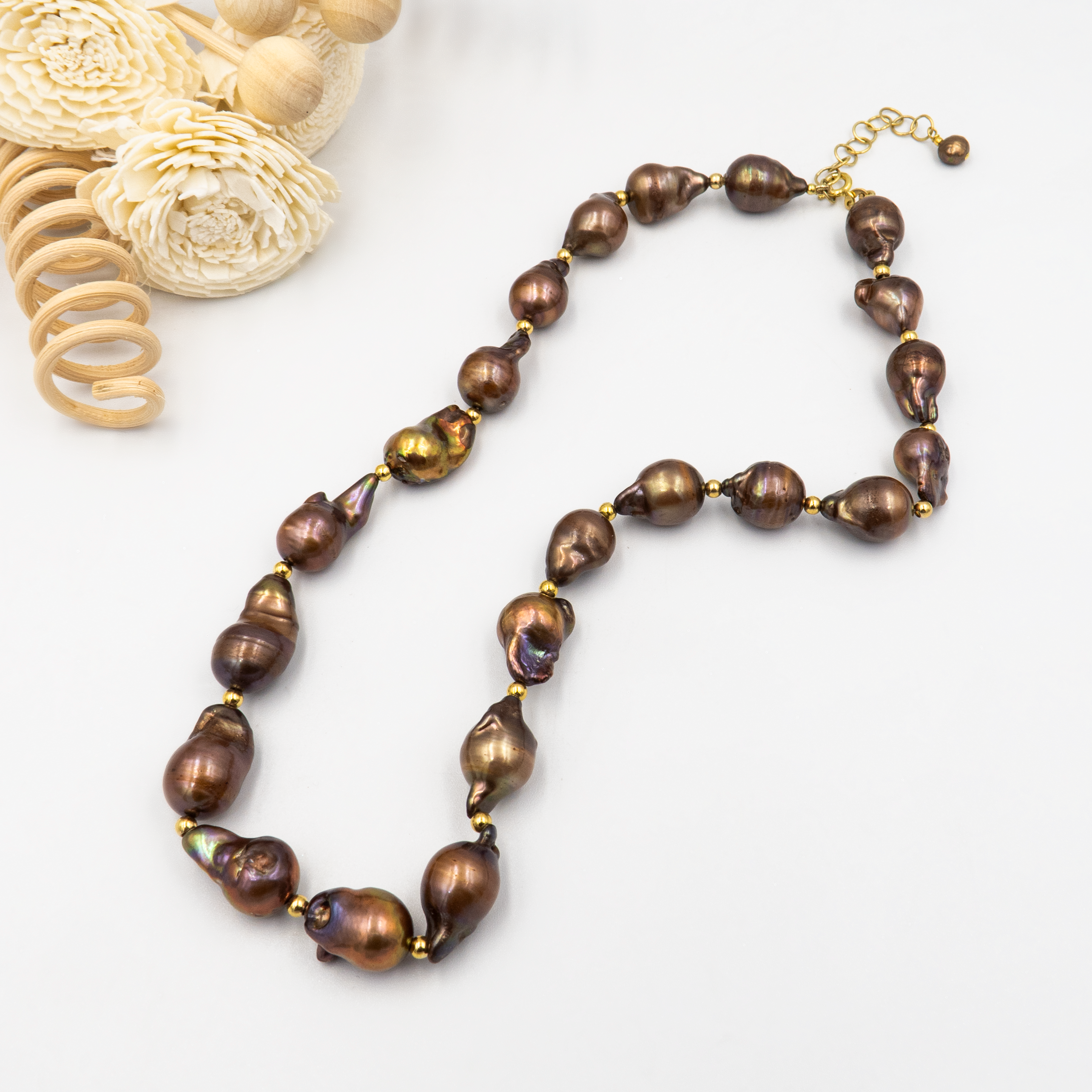 פנינים נדירות בעיצוב שמושך את העין המורכבת מפניני בארוק טבעיות בגוון שוקולד עמוק, עם אביזרים בציפוי זהב שמדגישים את צורתן הבלתי אחידה והמרקם החי של כל פנינה. הברק המטאלי החם של הפנינים משתלב באופן הרמוני עם הנגיעות המוזהבות, ויוצר רוך פראי בשילוב עם אלגנטיות חמה. שילוב ייחודי של פראיות מעודנת עם תחכום עיצובי – אידיאלי לאירוע ערב, להופעה עם אמירה או כמתנה מרשימה לאישה מיוחדת. פניני הבארוק, כל אחת עם אישיות משלה, הופכות את השרשרת הזו לתכשיט שהוא גם אמירה וגם תחושת חיבור אישי.