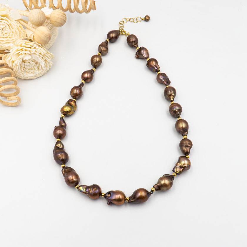 שרשרת יוקרתית בעלת מראה גולמי המורכבת מפניני בארוק טבעיות בגוון שוקולד עמוק, עם אביזרים בציפוי זהב שמדגישים את צורתן הבלתי אחידה והמרקם החי של כל פנינה. הברק המטאלי החם של הפנינים משתלב באופן הרמוני עם הנגיעות המוזהבות, ויוצר עיצוב שמביע שקט פנימי ועצמה שקטה. קו עיצובי טבעי עם קריצה לאופנה על־זמנית – אידיאלי לאירוע ערב, להופעה עם אמירה או כמתנה מרשימה לאישה מיוחדת. פניני הבארוק, כל אחת עם אישיות משלה, הופכות את השרשרת הזו לתכשיט שהוא גם אמירה וגם תחושת חיבור אישי.