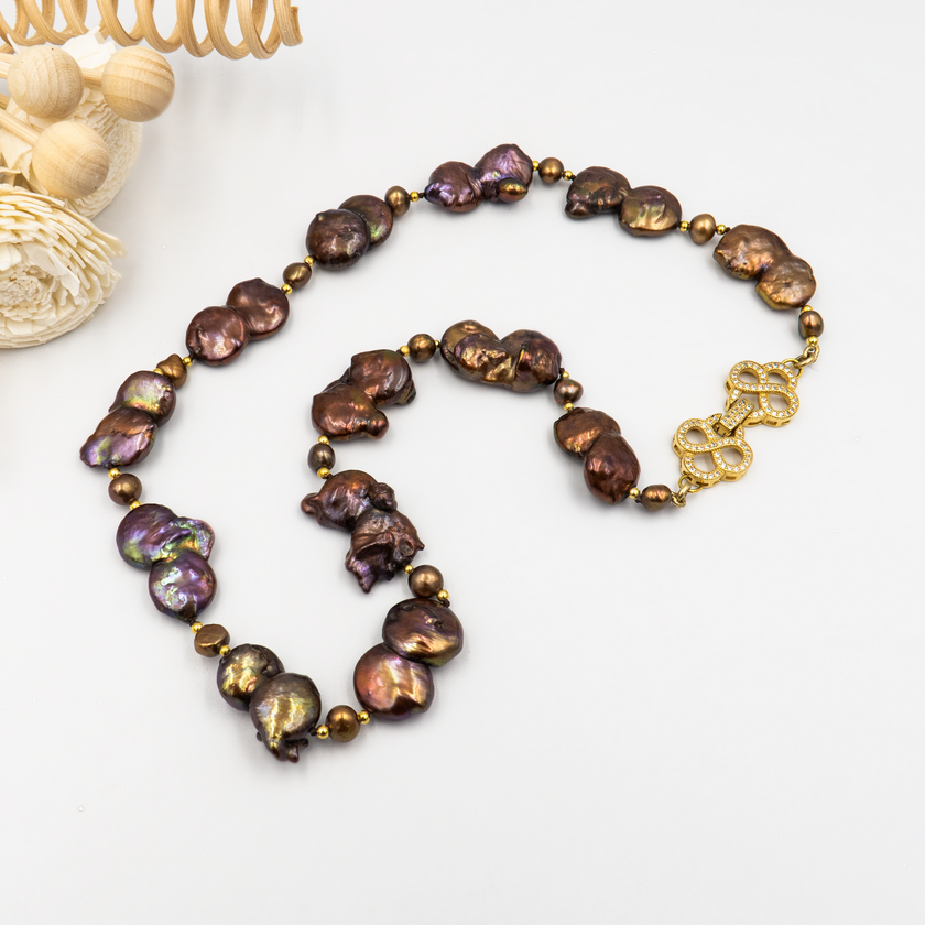 תכשיט בולט עם אופי טבעי המורכבת מפניני בארוק טבעיות בגוון שוקולד עמוק, עם אביזרים בציפוי זהב שמדגישים את צורתן הבלתי אחידה והמרקם החי של כל פנינה. הברק המטאלי החם של הפנינים משתלב באופן הרמוני עם הנגיעות המוזהבות, ויוצר תחושה של חומר חי ונושם. שילוב קלאסי ונועז כאחד – אידיאלי לאירוע ערב, להופעה עם אמירה או כמתנה מרשימה לאישה מיוחדת. פניני הבארוק, כל אחת עם אישיות משלה, הופכות את השרשרת הזו לשרשרת שלא דומה לשום דבר אחר – טבעית, אותנטית ומרשימה.