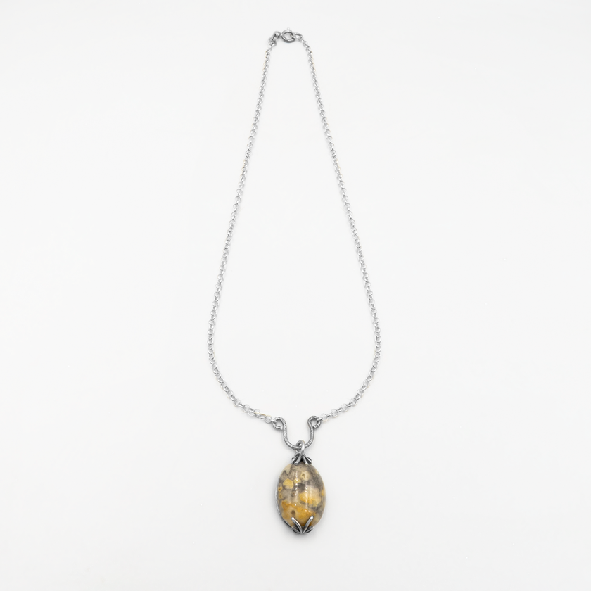 שרשרת ייחודית שמשלבת בין כסף בגימור עתיק לאבן טבעית במראה בלתי צפוי – גווני צהוב-חרדל משתלבים באפור עמוק עם נגיעות חום, ויוצרים דפוסים גיאולוגיים כמו ציור של הטבע עצמו. התליון, בצורת ביצה, מוחזק בשיבוץ פתוח המדגיש את האבן ולא מסתיר אותה, עם אלמנטים מתכתיים גולמיים בסגנון אורגני. זהו תכשיט שמספר סיפור של אדמה, עונות וטקסטורות.