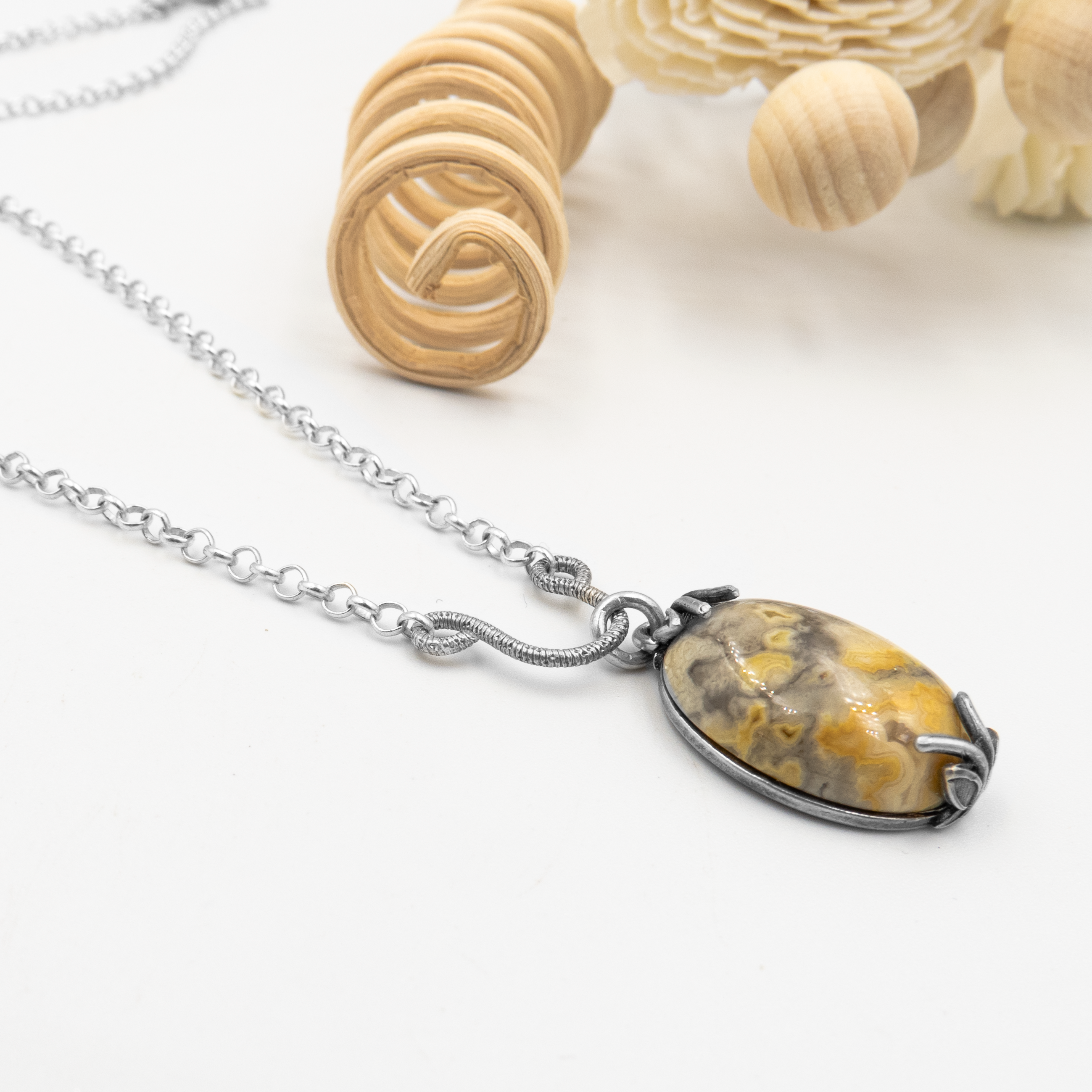 שרשרת בעבודת יד עם תליון אובלי עשוי אבן חן טבעית בגווני אדמה: כתום-צהוב, אפור וחום מעורבבים בציורי טבע מרהיבים. האבן המלוטשת משתלבת בתוך שיבוץ פתוח ממתכת כהה, המעוצבת כטפרים מעוגלים עם קווים דמויי ענפים או שורשים. שרשרת הכסף עצמה עדינה, בגימור מבריק למחצה, יוצרת ניגוד מרהיב מול גווני האבן. עיצוב שמאזן בין פראיות טבעית לאלגנטיות שקטה – מושלם לאוהבי אבנים לא שגרתיות.