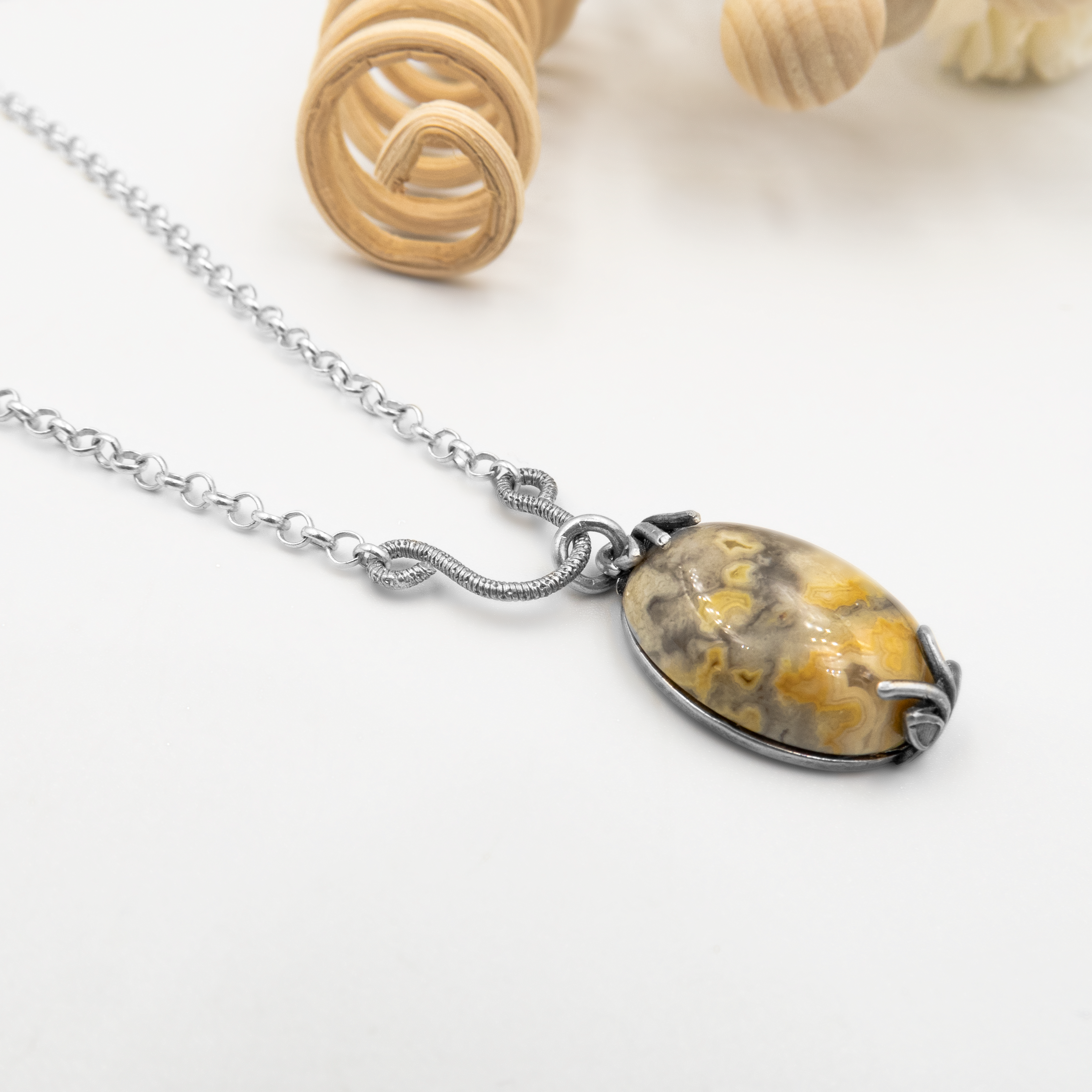 שרשרת עדינה בעלת תליון אובלי גדול, עשוי אבן טבעית בגוונים חמים של חום, אפור וצהוב זהוב. האבן יושבת בתוך שיבוץ פתוח בצורת טפרים גולמיים ממתכת כהה, המעוצבים בקווים זורמים ודינמיים, המדמים תנועה של שורשים או להבה. עיצוב שמתאים למי שמחפש פריט עם אנרגיה טבעית, גולמית ומעוררת השראה. התליון מושך את העין אך שומר על תחושת הרמוניה רגועה.