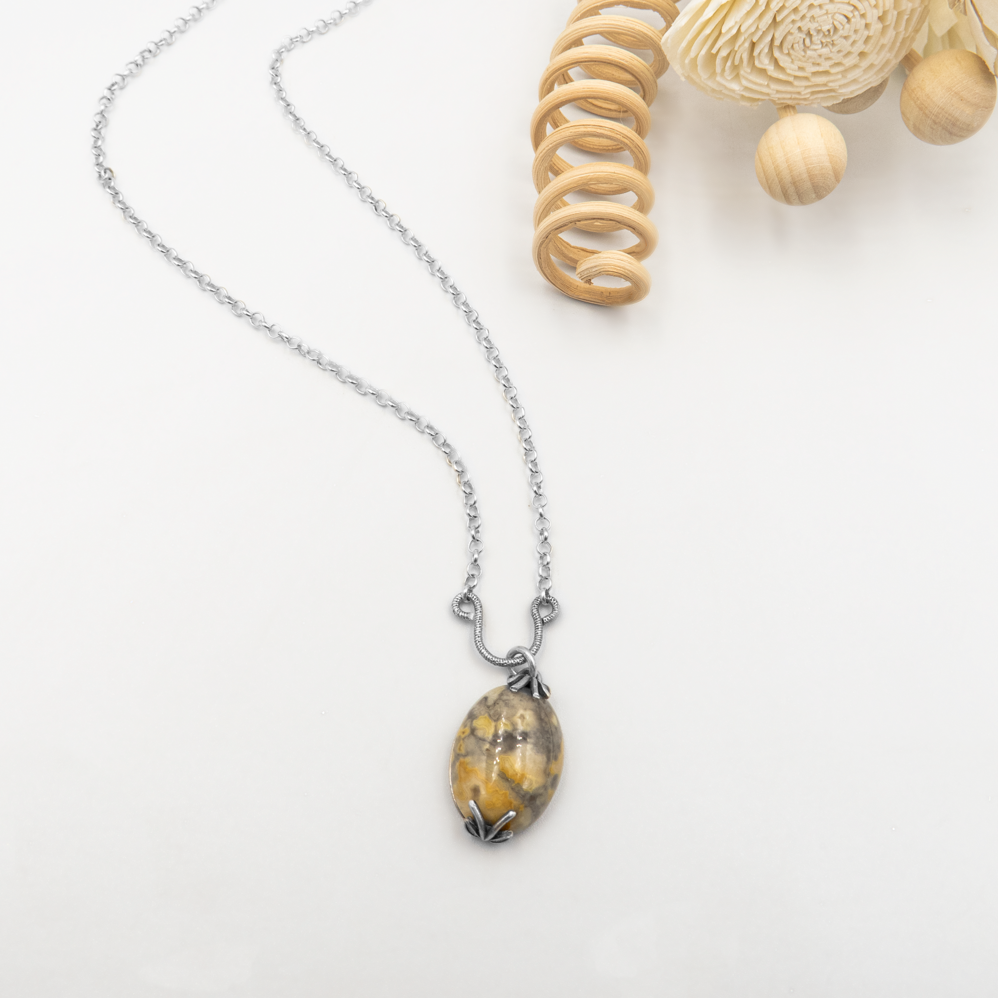 שרשרת כסף אלגנטית עם תליון אובלי מרשים עשוי אבן טבעית – ככל הנראה ג’ספר – בגוון זהב שרוף, אפור-כחלחל וחום מעושן. האבן המלוטשת מציגה מרקם מרהיב של שכבות טבעיות, וכמו נושאת בתוכה נוף הררי רחוק. המסגרת המתכתית המעוטרת מחזיקה אותה בארבע נקודות פתוחות, בסגנון אר נובו עדין, ומוסיפה לתכשיט תחושה של עתיקות וזמן. מתאים למי שמתחבר לטבע ולעיצוב שיש בו סיפור.