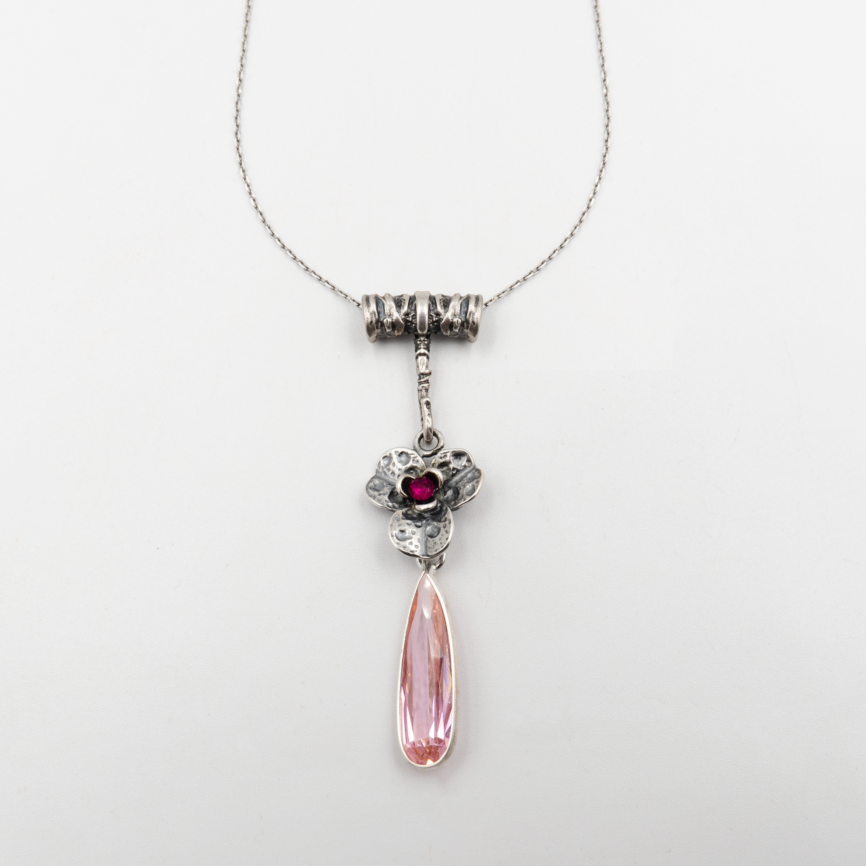 שרשרת כסף מעוצבת בעבודת יד עם תליון מרהיב, המשלב פרח בסגנון עתיק ועלי כותרת מעוטרים בפרטים עדינים. במרכז הפרח שובץ אבן ורודה עגולה בגוון עמוק, המדמה לב או נשיקה סודית. מתחתיו משתלשלת אבן טיפה ארוכה בגוון ורוד בהיר ונוצץ, המזכירה דמעה של פיה או טיפת שחר רכה. השרשרת מביאה עמה תחושה של רומנטיקה ונשיות עדינה, עם ניחוח של וינטג' וסיפור מהאגדות. מתאימה לאירועים חגיגיים, מתנה רומנטית או תוספת מרשימה ללוק יומיומי עם טוויסט פיוטי וייחודי.

