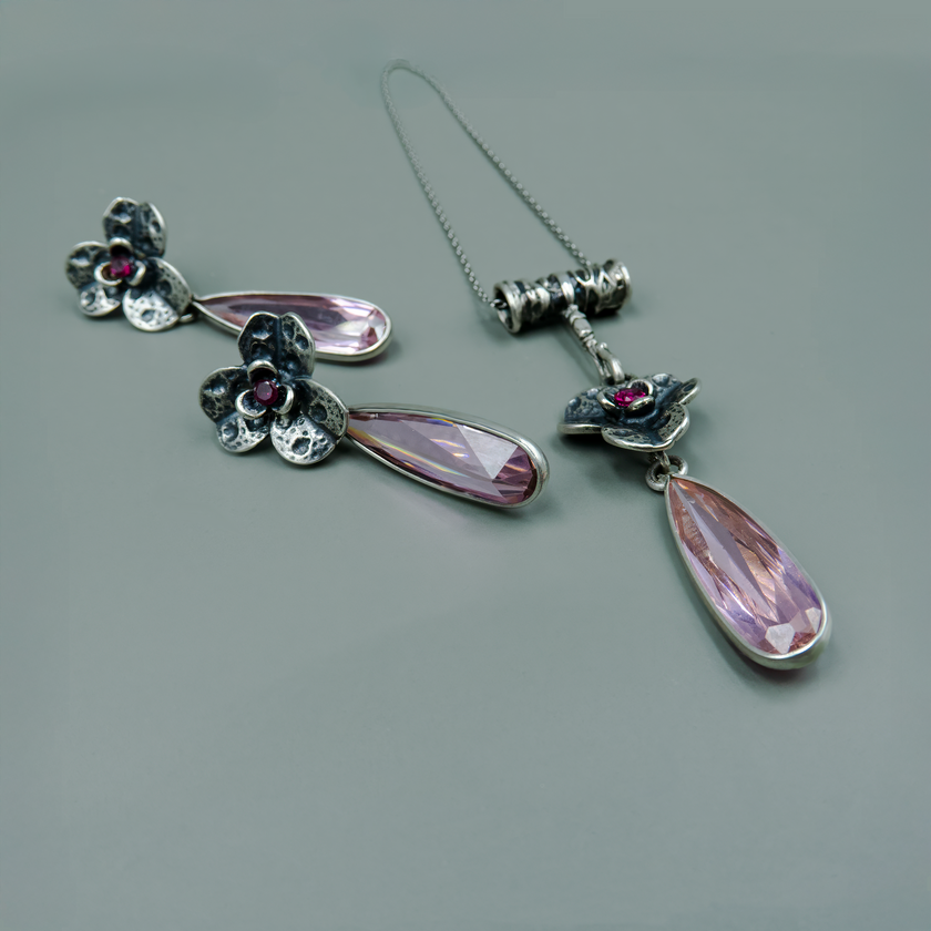 תליון מעוצב בהשראת עולם הפרחים והאגדות, עם אלמנטים כסופים מגולפים בעבודת יד ואבן מרכזית בגוון יין עמוק. מתחת לפרח משתלשלת טיפה ארוכה מבריקה בגוון ורדרד־שמפניה, המקנה לתכשיט תנועה חופשית ואלגנטית. השרשרת נראית כמו פריט מתוך קופסת תכשיטים נסתרת של נסיכה, ומשדרת סגנון אישי, רומנטי ונשי. הבחירה המושלמת כמתנה עם רגש.

העגילים להמחשה בלבד