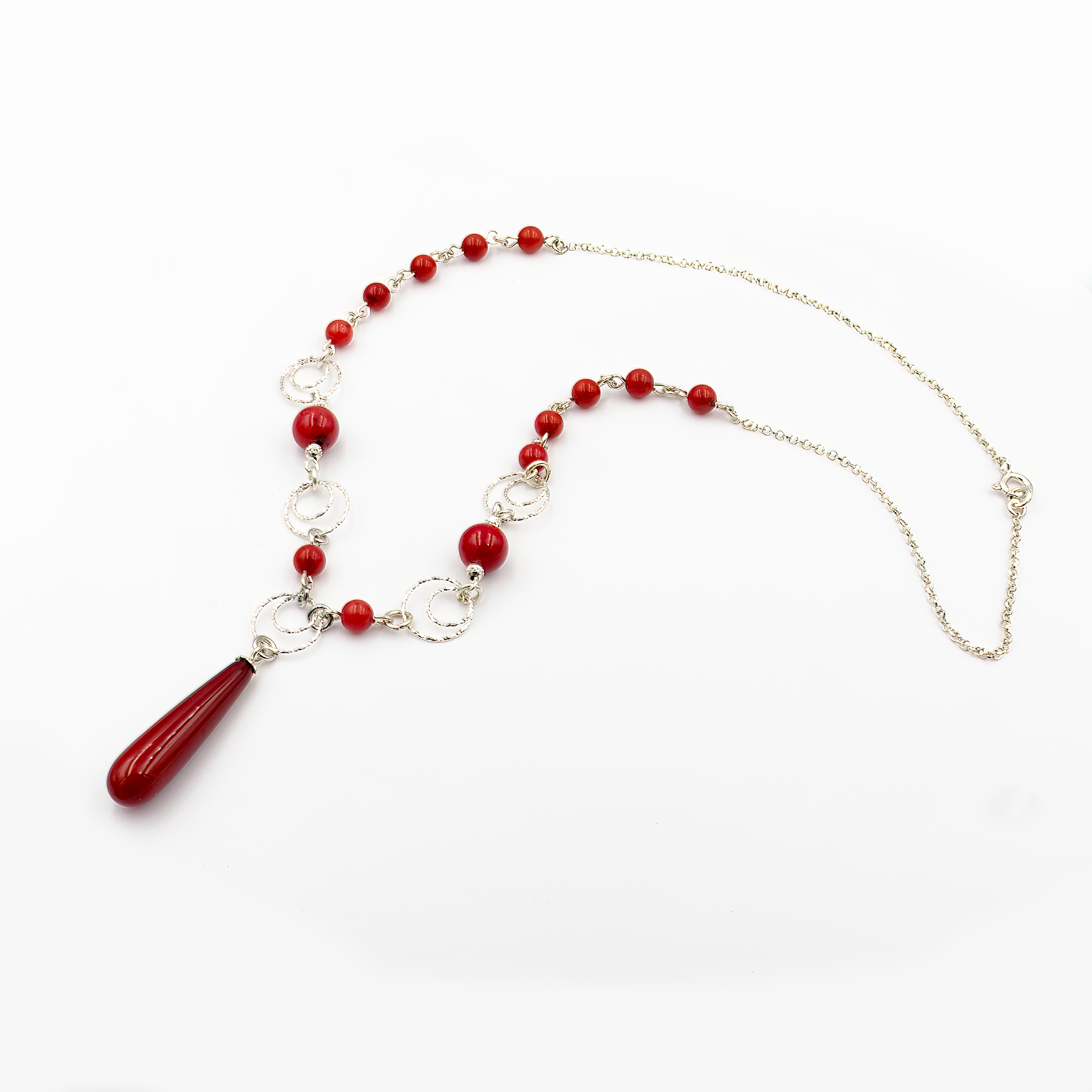צילום תקריב של שרשרת בעיצוב נועז עם חרוזים באדום עז, עשויים אלמוג סינתטי מבריק, בשילוב טבעות כסף מעוטרות. במרכז תלוי תליון ארוך בצורת טיפה באותו גוון. מוצגת על צוואר של דוגמנית עם ג'ינס, המדגיש את הניגוד בין הגוון האדום לבהיר.
