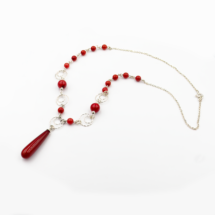 צילום תקריב של שרשרת בעיצוב נועז עם חרוזים באדום עז, עשויים אלמוג סינתטי מבריק, בשילוב טבעות כסף מעוטרות. במרכז תלוי תליון ארוך בצורת טיפה באותו גוון. מוצגת על צוואר של דוגמנית עם ג'ינס, המדגיש את הניגוד בין הגוון האדום לבהיר.