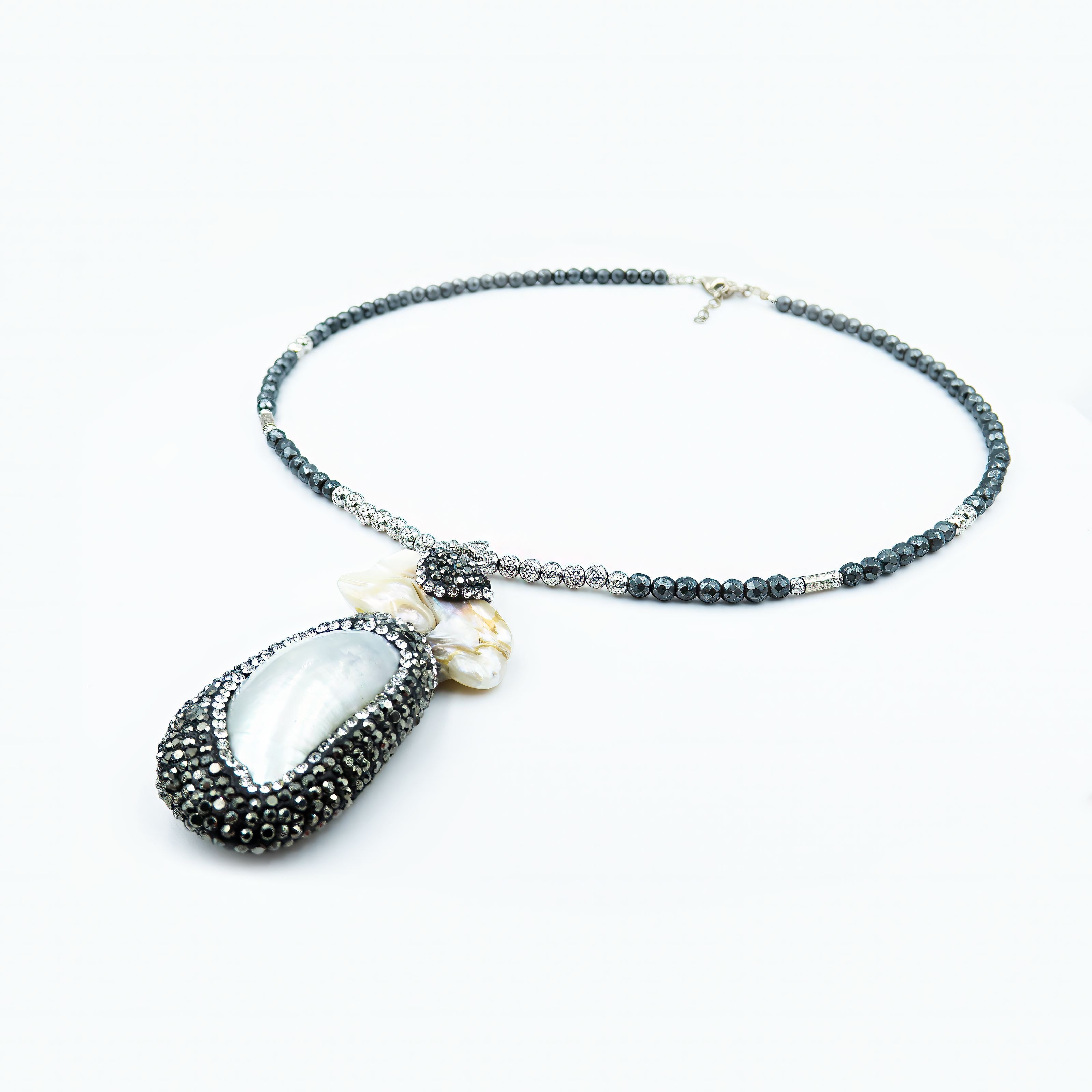 שרשרת אלגנטית בשחור ולבן עם תליון גדול בצורת טיפה משובץ אם הפנינה, עטוף גבול מנצנץ מקריסטלים שחורים. החרוזים לאורך השרשרת בגווני שחור מטאלי וכסף מבריקים, עם סגירה כסופה. שילוב מושלם בין גלאם מודרני לטבע ימי.

