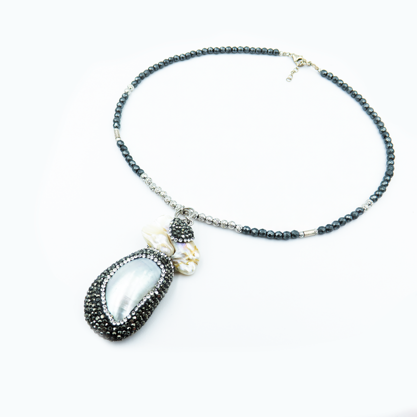 כמו אוצר מהים – תליון ייחודי עשוי צדף לבן שנראה כאילו נשטף הרגע אל החוף, עטוף באלפי קריסטלים שחורים נוצצים. השרשרת משלבת חרוזים כהים בגווני המעמקים, ומשדרת תחושת עומק, מסתורין ויוקרה טבעית.