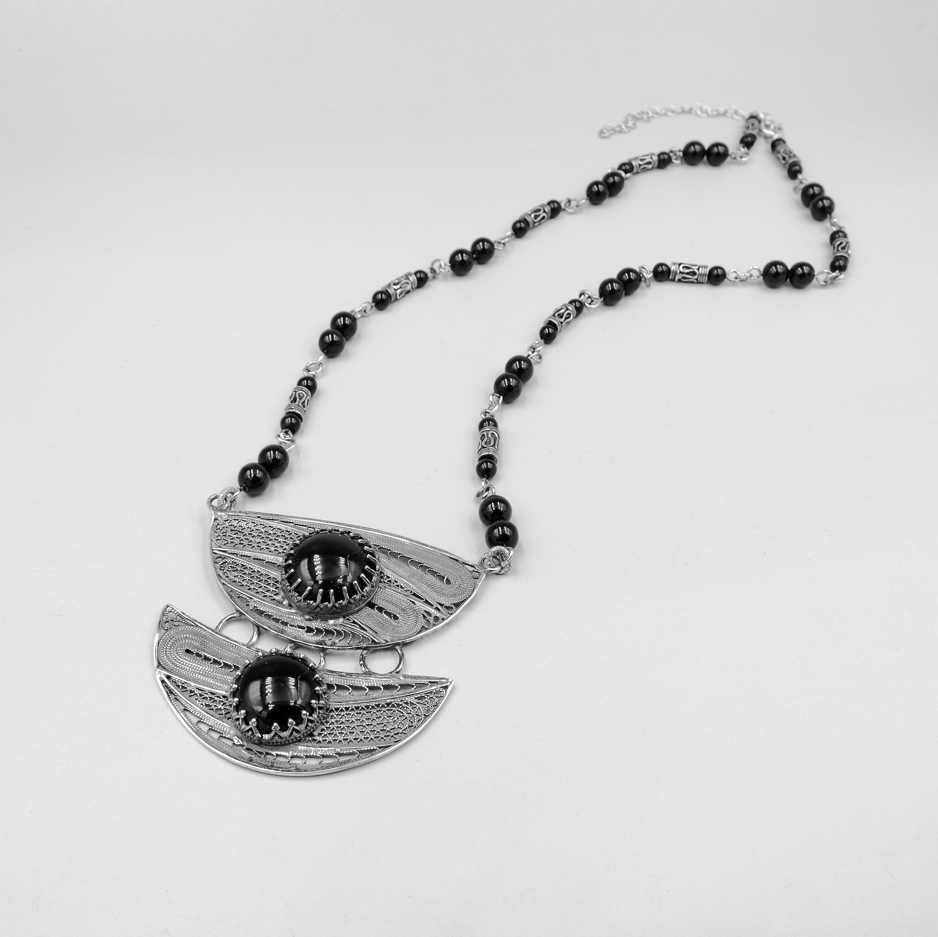 שרשרת כסף עם עיצוב גאומטרי ייחודי, המשלבת תליוני כסף בעבודת פיליגרין מסורתית, עם אבני אוניקס שחורות במרכזם. השרשרת מורכבת מאלמנטים אתניים, חרוזים מעוטרים בעבודת יד ואוניקס מבריק, ליצירת נוכחות אמנותית שמוסיפה עניין לכל הופעה. עיצוב שמחבר בין שורשיות מסורתית לבין קווים מודרניים, מתאים לאישה שאוהבת תכשיטים עם אופי ונוכחות. החומרים האיכותיים וההרכב האמנותי מעניקים לשרשרת תחושת חיבור לשורשים עם קריצה עכשווית.