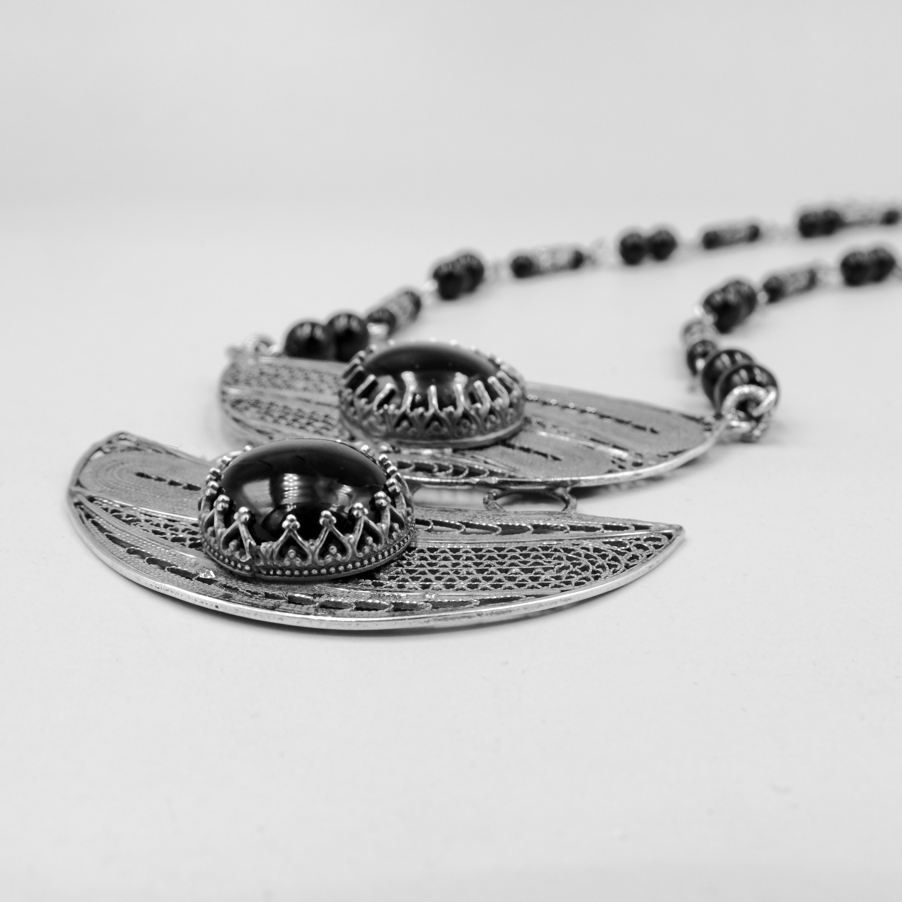 שרשרת כסף עם עיצוב גאומטרי ייחודי, המשלבת שני תליוני כסף עם עיטורים עדינים, עם אבני אוניקס שחורות במרכזם. השרשרת מורכבת מאלמנטים אתניים, חרוזים מעוטרים בעבודת יד ואוניקס מבריק, ליצירת נוכחות אמנותית שמוסיפה עניין לכל הופעה. עיצוב שמחבר בין שורשיות מסורתית לבין קווים מודרניים, מתאים לאישה שאוהבת תכשיטים עם אופי ונוכחות. החומרים האיכותיים וההרכב האמנותי מעניקים לשרשרת איזון בין יוקרה שקטה לבין עיצוב מלא תוכן.