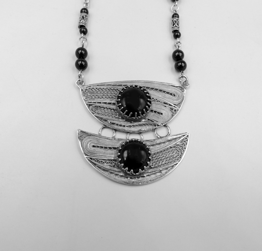 שרשרת כסף עם עיצוב גאומטרי ייחודי, המשלבת שני תליוני כסף עם עיטורים עדינים, עם אבני אוניקס שחורות במרכזם. השרשרת מורכבת מאלמנטים אתניים, חרוזים מעוטרים בעבודת יד ואוניקס מבריק, ליצירת מראה שמחבר בין אמנות ואלגנטיות. עיצוב שמחבר בין שורשיות מסורתית לבין קווים מודרניים, מתאים לאישה שאוהבת תכשיטים עם אופי ונוכחות. החומרים האיכותיים וההרכב האמנותי מעניקים לשרשרת איזון בין יוקרה שקטה לבין עיצוב מלא תוכן.