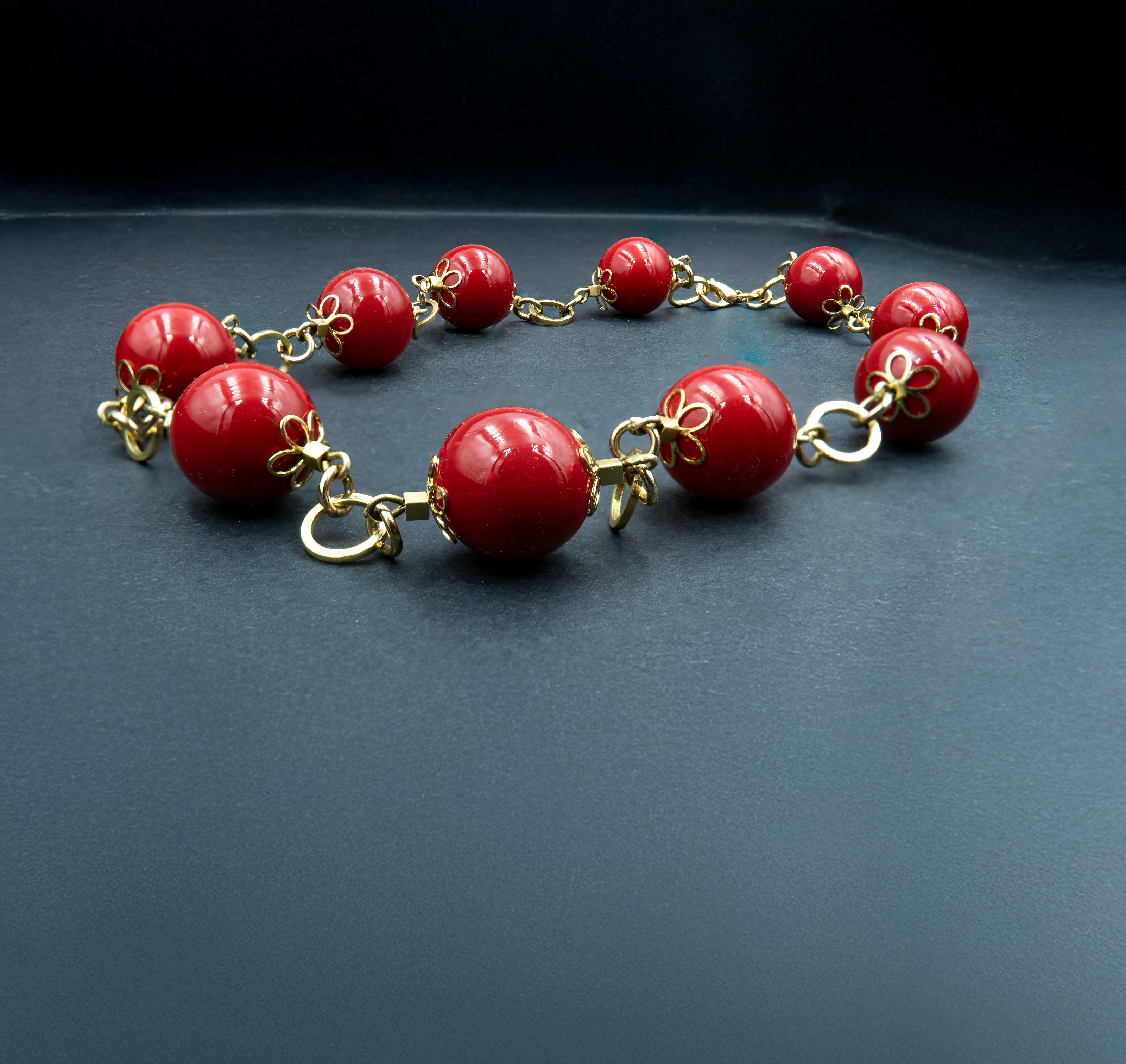 תמונה של שרשרת קלאסית בעיצוב דרמטי ובולט, עשויה חרוזים עגולים גדולים בגוון אדום עמוק ומבריק. כל חרוז מעוטר בסוגר מתכתי מוזהב בצורת פרח, המחבר אותו לחרוז הבא באמצעות חוליות מוזהבות עבות. רקע כהה ניטרלי מדגיש את הברק והצבע של החרוזים, ויוצר תחושת יוקרה ונוכחות.