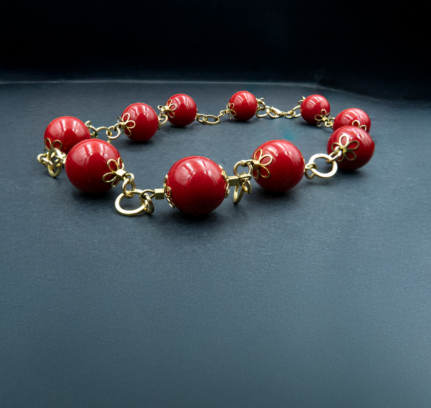 תמונה של שרשרת קלאסית בעיצוב דרמטי ובולט, עשויה חרוזים עגולים גדולים בגוון אדום עמוק ומבריק. כל חרוז מעוטר בסוגר מתכתי מוזהב בצורת פרח, המחבר אותו לחרוז הבא באמצעות חוליות מוזהבות עבות. רקע כהה ניטרלי מדגיש את הברק והצבע של החרוזים, ויוצר תחושת יוקרה ונוכחות.