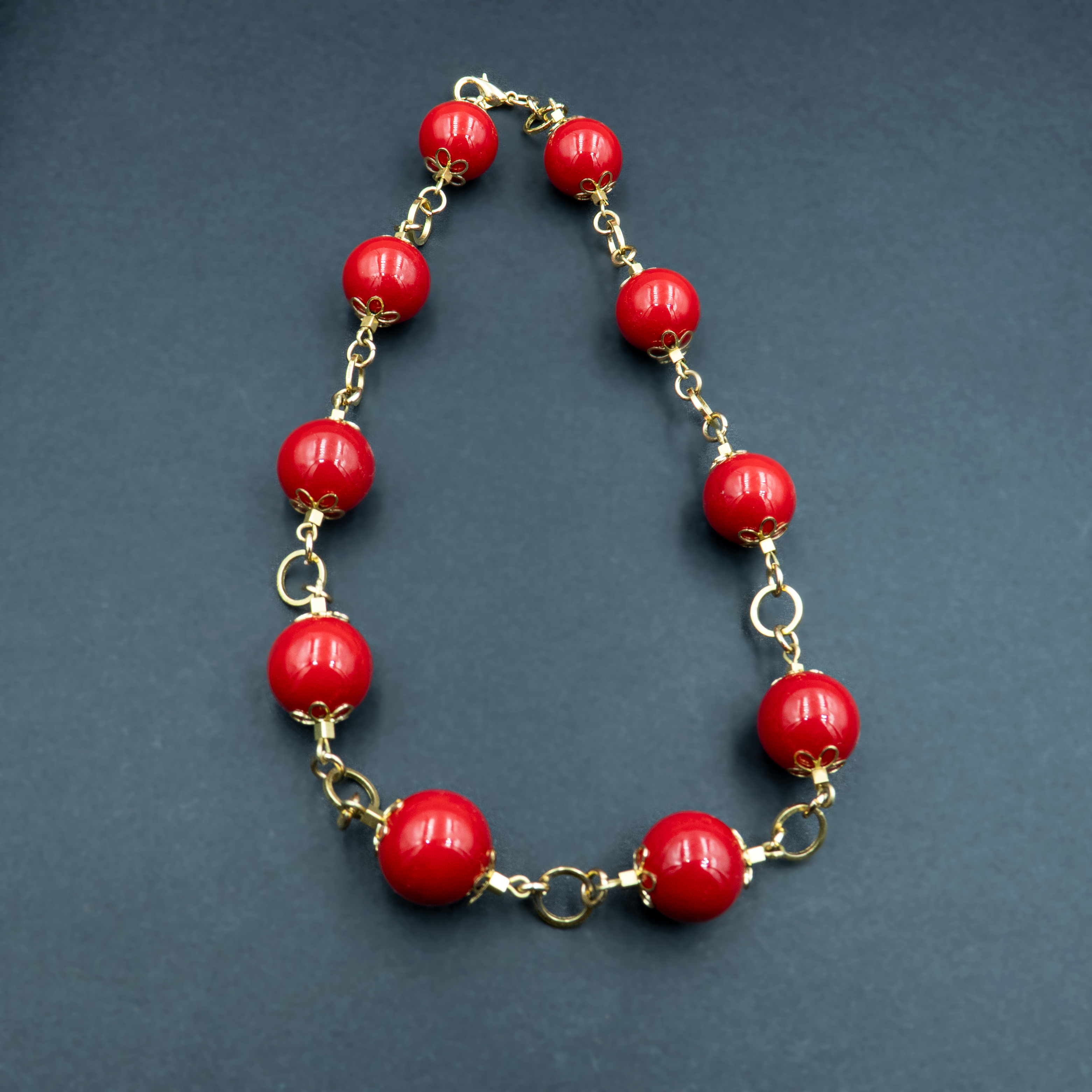 תכשיט בולט בעיצוב וינטג'־מודרני, הכולל חרוזים עגולים אדומים מבריקים בגודל משתנה, מחוברים זה לזה באמצעות אלמנטים מוזהבים עם עיטורים פרחוניים. הרקע הכהה מוסיף עומק ואלגנטיות, ומדגיש את הניגוד בין האדום העז לבין הזהב הנוצץ.