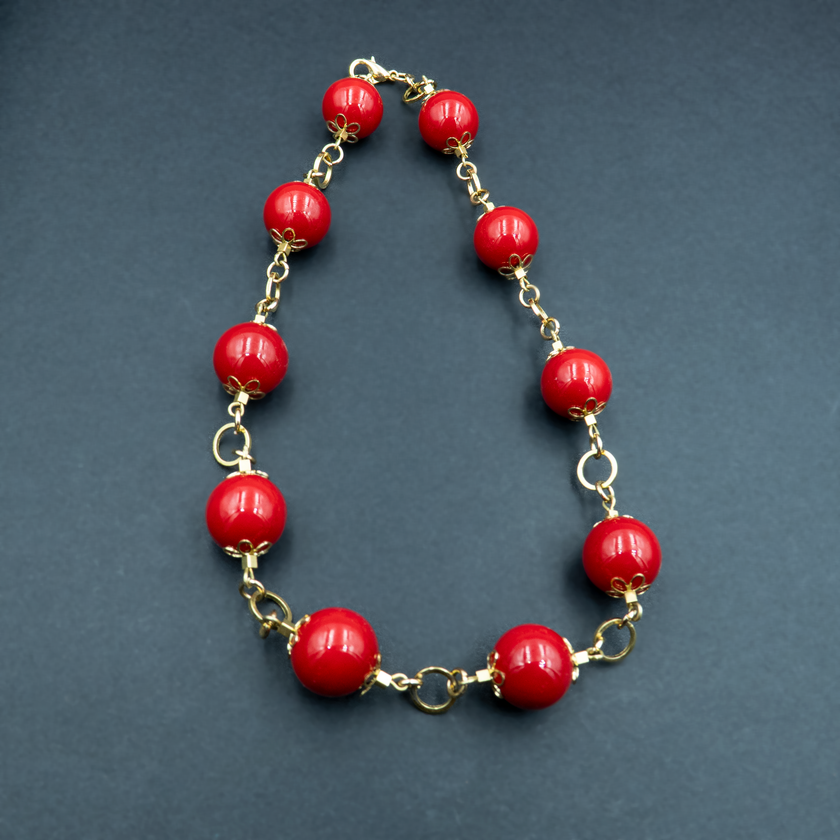 תכשיט בולט בעיצוב וינטג'־מודרני, הכולל חרוזים עגולים אדומים מבריקים בגודל משתנה, מחוברים זה לזה באמצעות אלמנטים מוזהבים עם עיטורים פרחוניים. הרקע הכהה מוסיף עומק ואלגנטיות, ומדגיש את הניגוד בין האדום העז לבין הזהב הנוצץ.