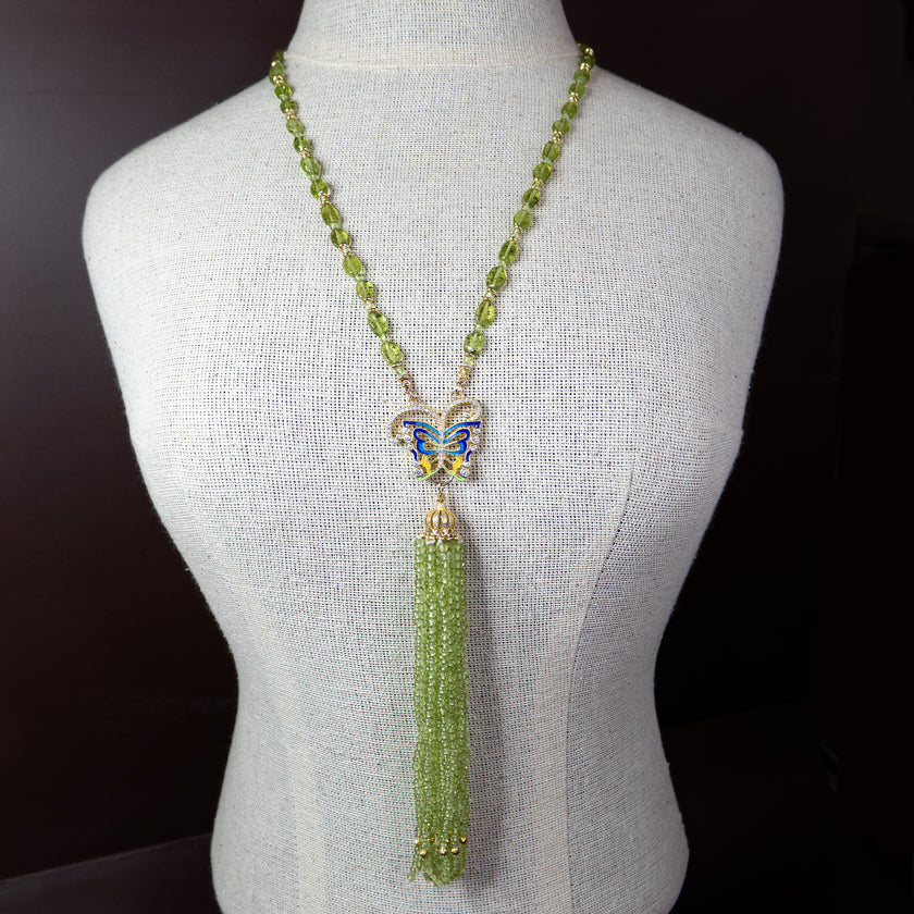 שרשרת ארוכה המעוצבת מחרוזים מלוטשים בגוון ירוק פרידוט, בגדלים משתנים. שיא התכשיט הוא תליון פרפר בגימור מוזהב, המעוטר באמייל בגוני כחול, לבן וזהב, שממנו משתלשלים כעשרה גדילים צפופים של חרוזים קטנטנים בצורת טיפה, היוצרים תנועה מרהיבה עם כל תזוזה. האלמנטים מחוברים בעזרת חבקי זהב דקורטיביים והשרשרת נסגרת באמצעות סוגר לובסטר. הרקע בתמונה בגוון ורוד פודרה שמחמיא לצבעי התכשיט ומדגיש את תחושת הרוך והאלגנטיות.