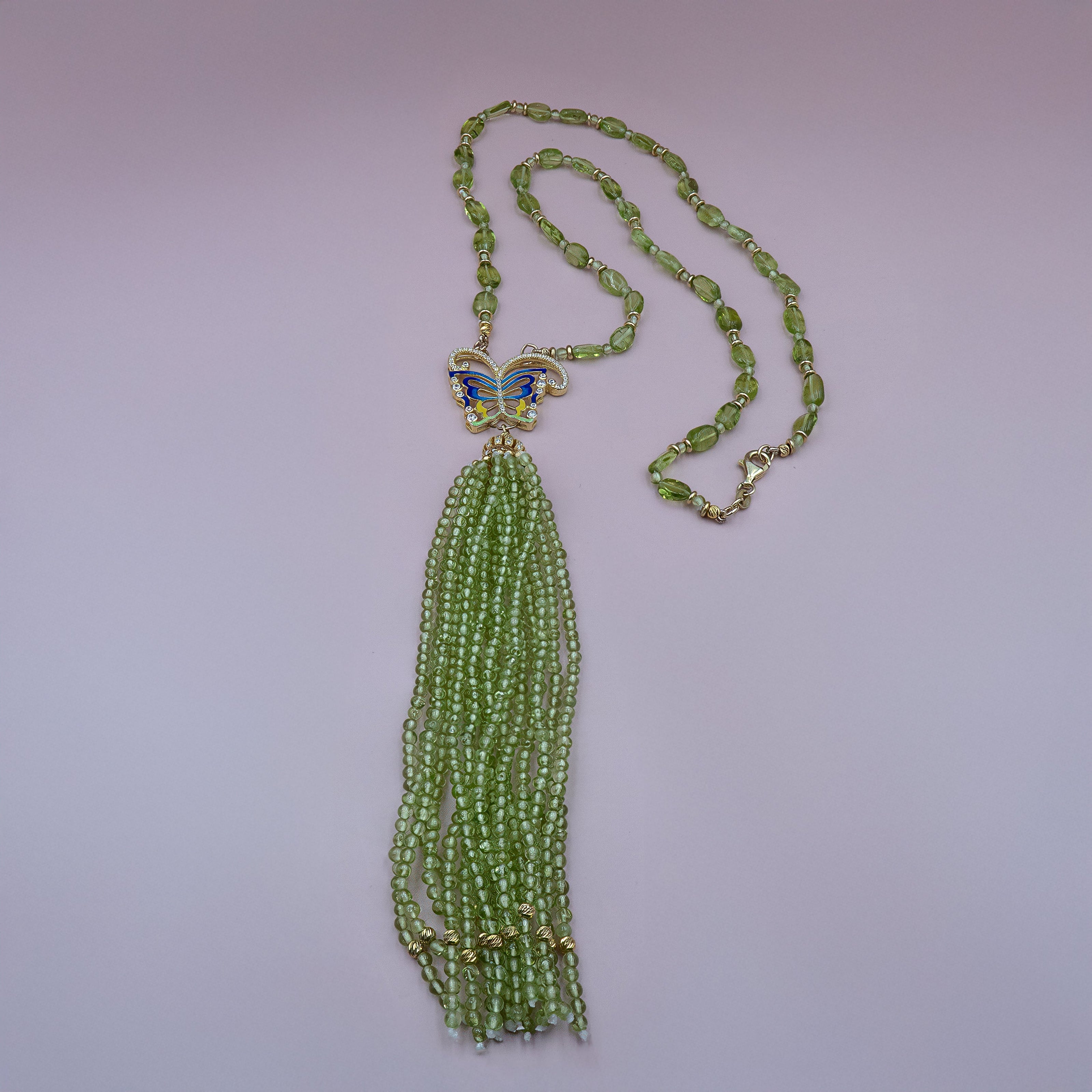 שרשרת בעבודת יד המשלבת חרוזים ירקרקים עשויים מאבן פרידוט טבעית בגוונים רכים, מסודרים בחריזה מדורגת ויוקרתית. במרכז השרשרת תליון ייחודי בצורת פרפר מוזהב, משובץ באמייל כחול ולבן ונצנוצים קטנים. מהפרפר משתלשלים כ־10 גדילים צפופים של חרוזי פרידוט קטנים היוצרים תנועה חופשית ועשירה. השרשרת נסגרת בסוגר לובסטר מוזהב ומעוצבת בקו קליל אך מלא נוכחות – פריט שמושך את העין ומוסיף צבע ותנועה לכל לוק.