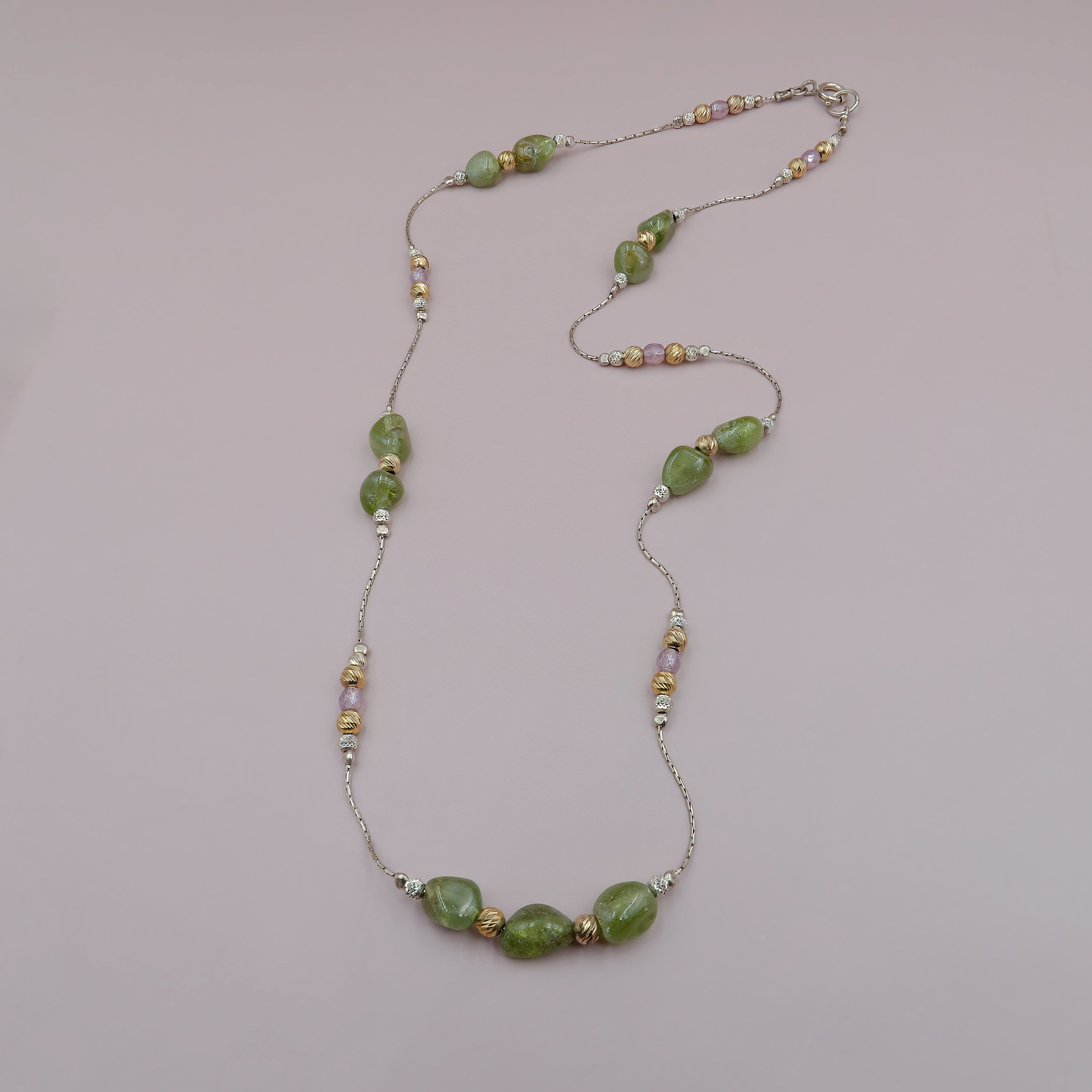 תכשיט בעיצוב נשי ורך, משלב אבנים ירוקות גולמיות עם חרוזים מתכתיים מעוטרים בגוונים של כסף, זהב וורוד מעושן. השרשרת עדינה אך בעלת נוכחות, ומתאימה להשלמת מראה עם ניחוח טבעי ורומנטי. הרקע הניטרלי מחמיא לצבעים הרכים ומדגיש את המרקמים המשתנים של החרוזים.
