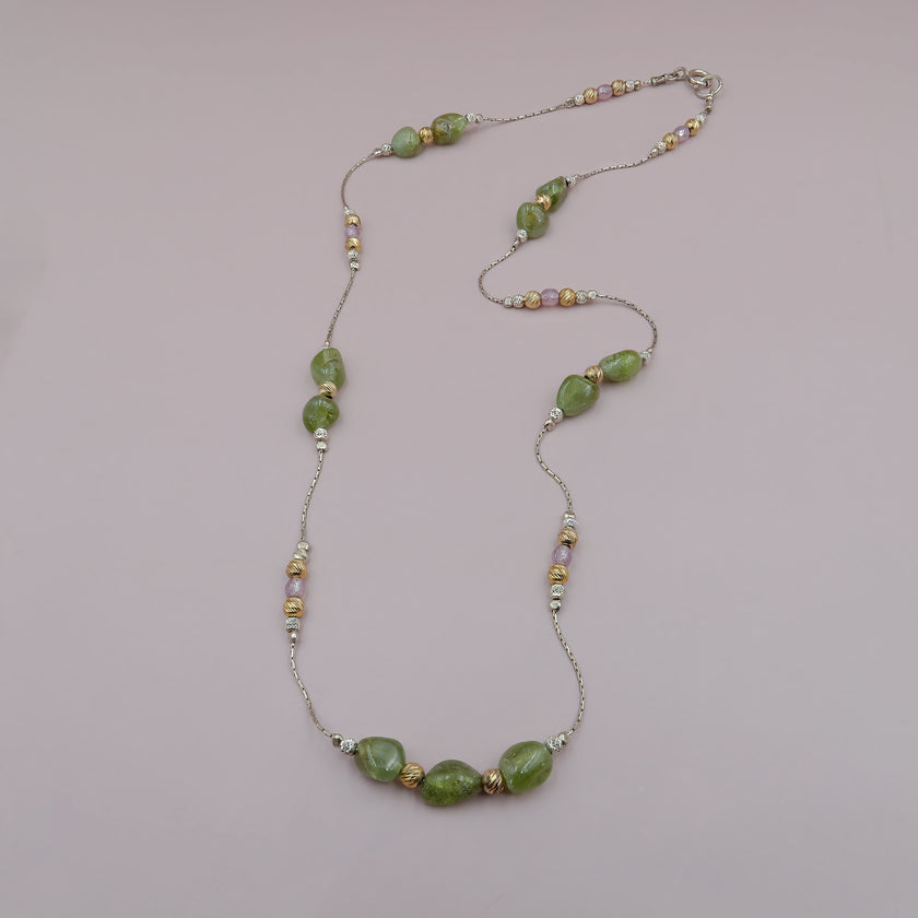 תכשיט בעיצוב נשי ורך, משלב אבנים ירוקות גולמיות עם חרוזים מתכתיים מעוטרים בגוונים של כסף, זהב וורוד מעושן. השרשרת עדינה אך בעלת נוכחות, ומתאימה להשלמת מראה עם ניחוח טבעי ורומנטי. הרקע הניטרלי מחמיא לצבעים הרכים ומדגיש את המרקמים המשתנים של החרוזים.