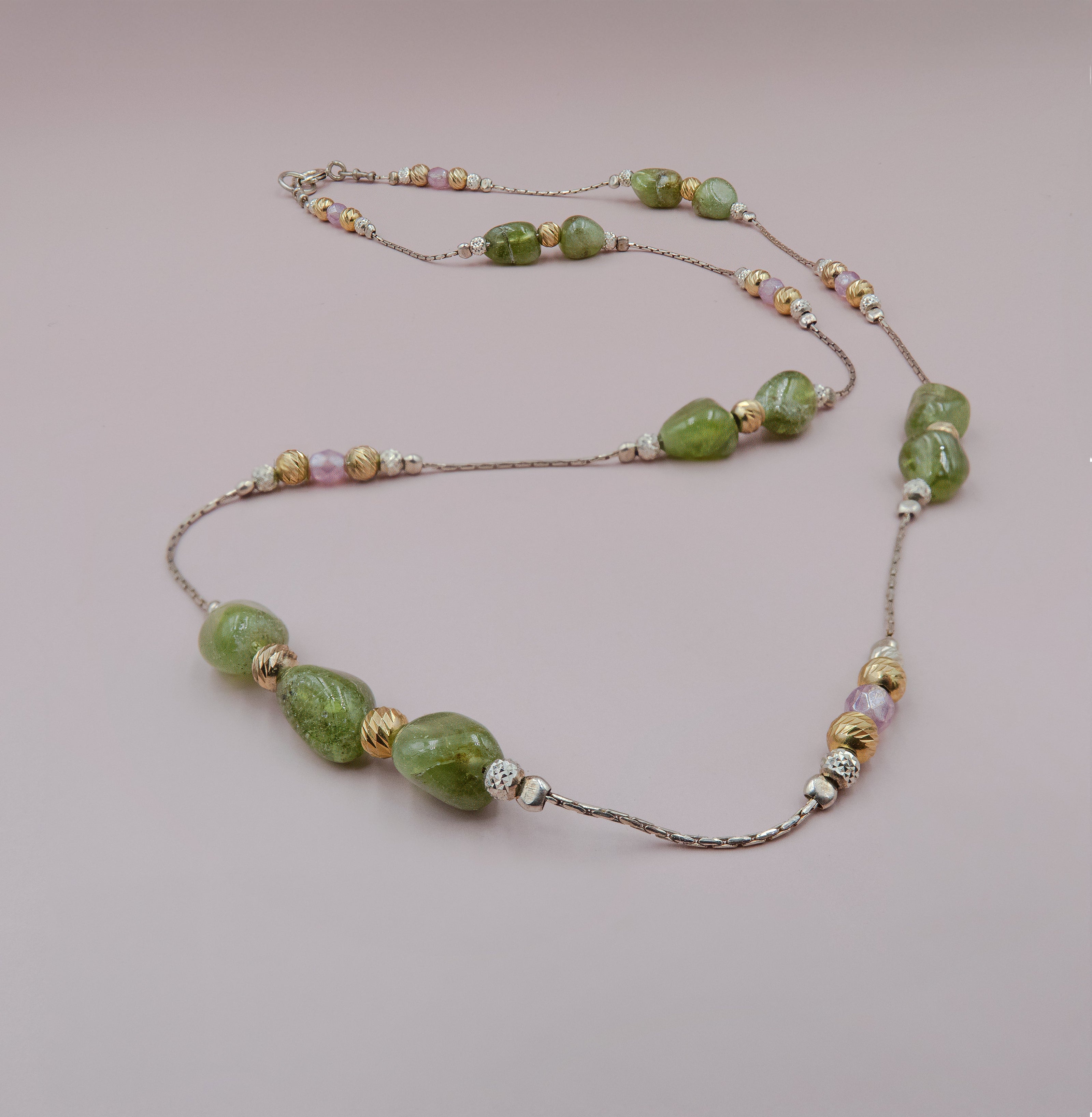 שרשרת אלגנטית עם חרוזים ירוקים בלתי אחידים, כנראה פרידוט או סרפנטין, משולבים עם חרוזים מוזהבים, כסופים וסגלגלים. השרשרת עשויה מחוט מתכתי עדין ומבריק, עם שילוב צבעים טבעי ורך המעניק לה מראה קלאסי ונשי. התמונה מצולמת על רקע ורוד בהיר ניטרלי, המדגיש את צבעי האבנים ואת הניצוץ המתכתי של החרוזים.

