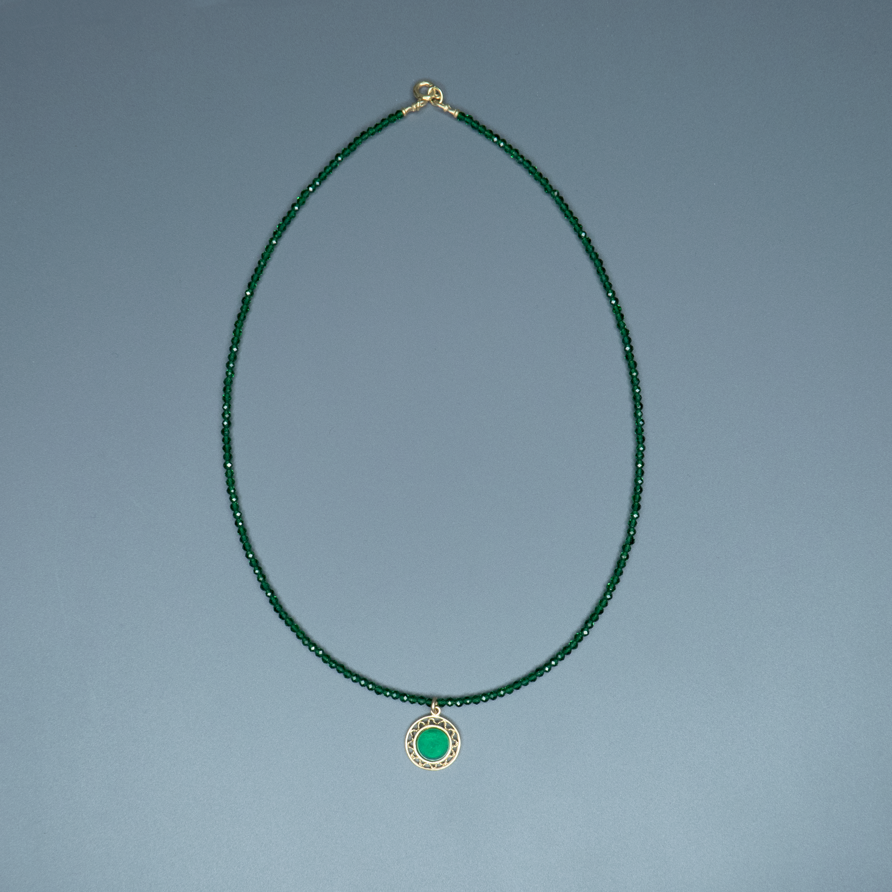 תכשיט שמזכיר יער שקט – קולר ירוק עשוי חרוזים זעירים ונוצצים שמתחברים לתליון עגול מוזהב עם אבן ירוקה בוהקת בלב ליבו. השילוב בין הזהב לירוק יוצר הרמוניה של טבע ואלגנטיות. צילום על צוואר חשוף עם חולצה לבנה, מדגיש את העדינות והנוכחות השקטה של התכשיט.