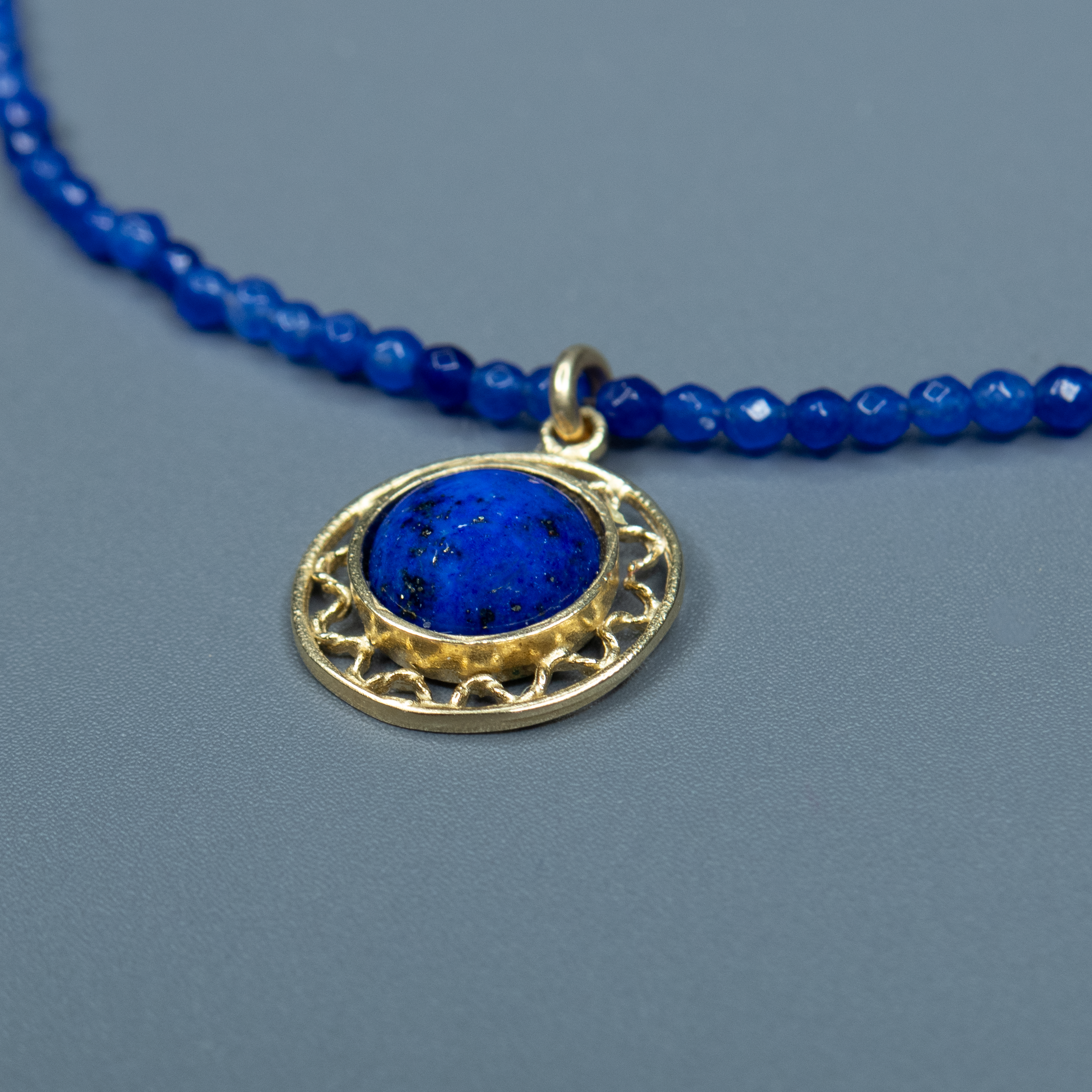 קולר אלגנטי בגווני כחול עמוק וזהב, עם תליון עגול המזכיר ירח כחול זורח בליל קיץ. האבן המרכזית הכחולה מהפנטת במרקם ובברק שלה, ומוקפת במסגרת מוזהבת עדינה עם חריטה דקורטיבית. השרשרת נצמדת בעדינות לצוואר ומוסיפה נגיעה של עומק ורוחניות למראה. תכשיט שמביא איתו תחושת מסתורין, השראה ורכות.