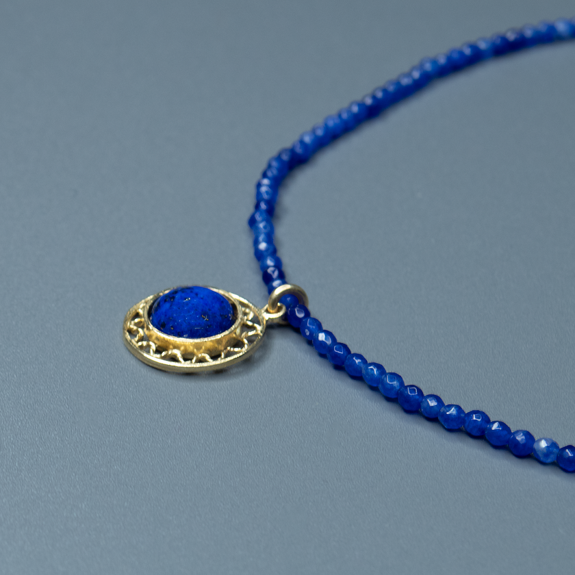 שרשרת קולר עדינה עשויה חרוזים כחולים כהים, כמו שמיים ליליים זרועים כוכבים, ובמרכזה תליון עגול מוזהב עם אבן כחולה בוהקת בלבו – דומה למראה הירח המלא מעל ים עמוק. העיצוב הקלאסי משתלב בהרמוניה עם האלמנטים הגאומטריים, ויוצר מראה של איזון והרמוניה. תכשיט מושלם למי שמבקשת להוסיף מגע של עומק שקט ואור פנימי לכל הופעה.