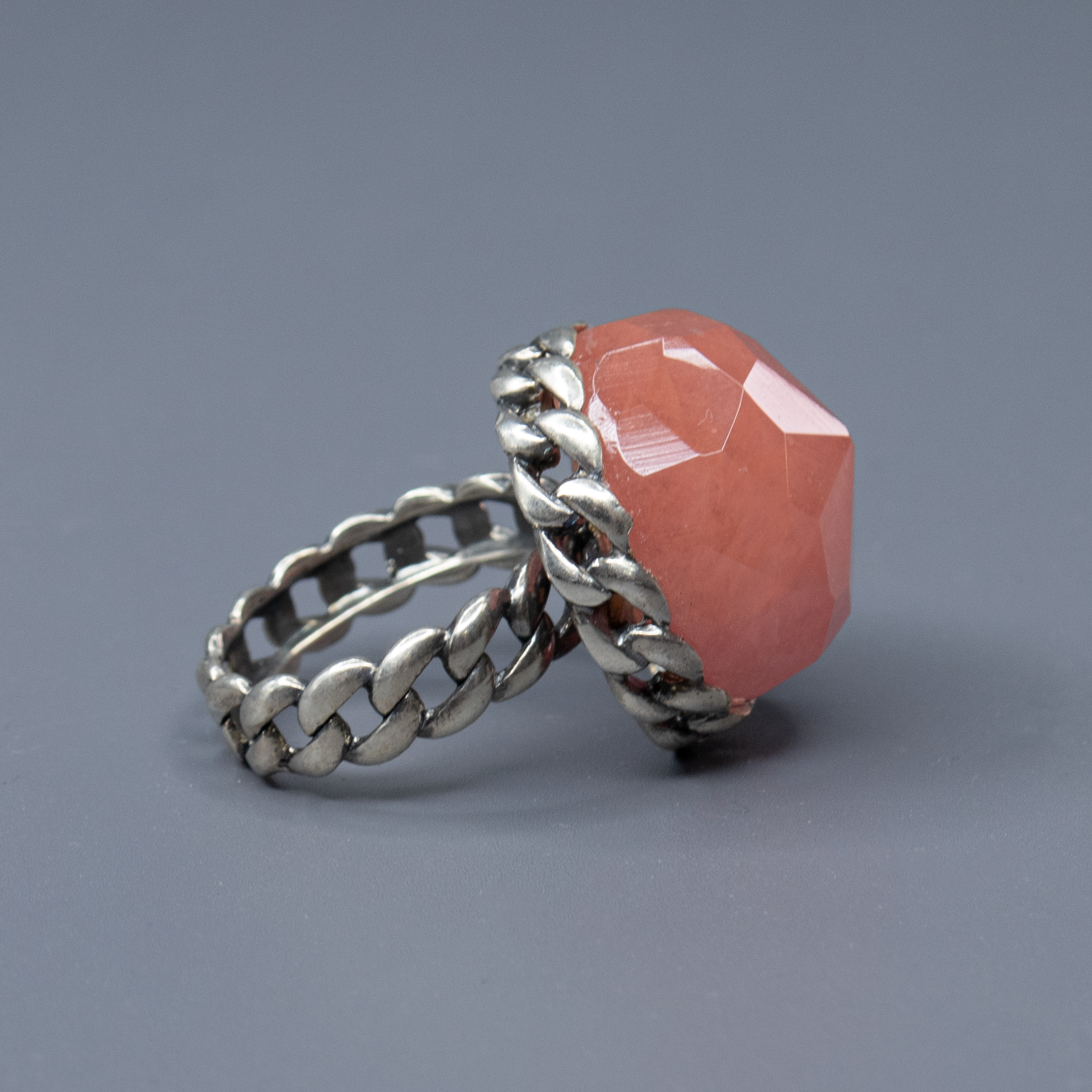 טבעת כסף דרמטית בהכנה קפדנית, מעוטרת באבן גדולה בגוון ורוד־קורל עם חיתוך פאות (faceted) שמדגיש את הברק והעומק של האבן. העיצוב משלב בסיס כסף בעבודת חריטה בדוגמה קלועה, המעניק לטבעת מראה חזק אך אלגנטי, שמזכיר טבעת שרשרת או סגנון בוהו וינטג' מתוחכם. האבן הגדולה הופכת את הטבעת לטבעת יוצאת דופן, כזה שלא עובר מתחת לרדאר, אך שומר על איזון אסתטי. גוון האבן מחמיא לעור, מתאים לקיץ ולחורף כאחד, ויוצר ניגוד יפהפה עם גוון המתכת. תכשיט מיוחד שמדגיש אישיות, טעם ייחודי ואמירה אישית לא שגרתית. מושלמת כטבעת מתנה, ליום־יום או