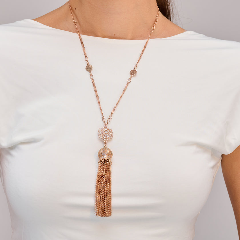 צילום תקריב של שרשרת ארוכה מעוצבת בגוון רוז גולד, עם פרח עדין ממתכת, כדור עגול מחורר בגימור תחרה, ותליון באורך מרובה שרשראות דקות. מונחת על בובת תצוגה מבד בהיר. ברקע נראית שקית יוקרתית עם לוגו המעידה על מותג מקומי מעוצב בקפידה. תכשיט עם אופי רומנטי-מודרני.