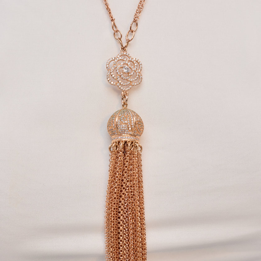 שרשרת נשית ארוכה בגוון זהב ורוד מבריק, עם תליון ורד מתכתי פתוח, עיטור כדורי וגדילים ארוכים משרשראות דקות ואווריריות. צילום באווירה אולפנית על בובת תצוגה נייטרלית, עם נגיעה מותגית ברקע. מושלם כתכשיט לאירועים או לשדרוג לוק יומיומי.