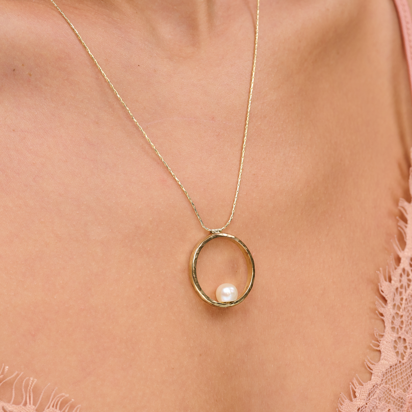 שרשרת בעיצוב אלגנטי עם תליון מוזהב בגודל דומיננטי, המשובץ בפנינה לבנה קלאסית עם ברק טבעי – תכשיט קלאסי בעל נוכחות בולטת. הפנינה ממוקמת במרכז התליון ומקרינה אור רך וטבעי, תוך יצירת ניגוד מחמיא עם הציפוי הזהוב. עיצוב השרשרת משלב בין עידון נשי עם נגיעה עכשווית, ומתאים לאירועים חגיגיים כמו גם ללבוש יומיומי עם נגיעה נשית. פנינים ידועות כמזוהות עם סטייל נשי טהור – ויחד עם הזהב, נוצרת הרמוניה מושלמת בין אלגנטיות לחמימות.