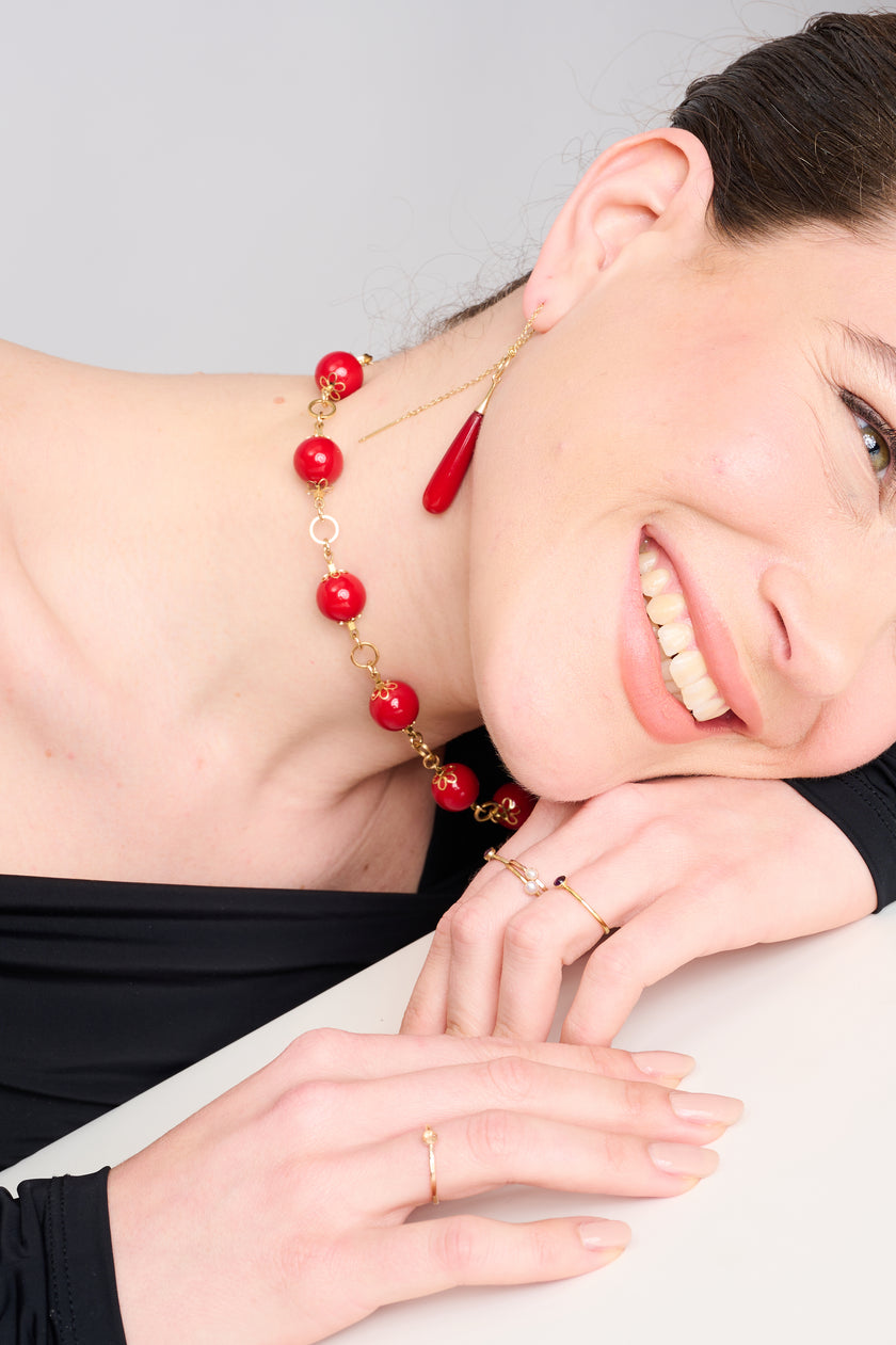 צילום תקריב של שרשרת עם חרוזים עגולים בגוון אדום בוהק, מחוברים בעבודת יד עם מתכת מוזהבת הכוללת עיטורים בצורת פרח. התכשיט שוכן על רקע כהה ומינימליסטי, המדגיש את הברק של הצבעים והעיצוב הקלאסי עם נגיעה מודרנית.