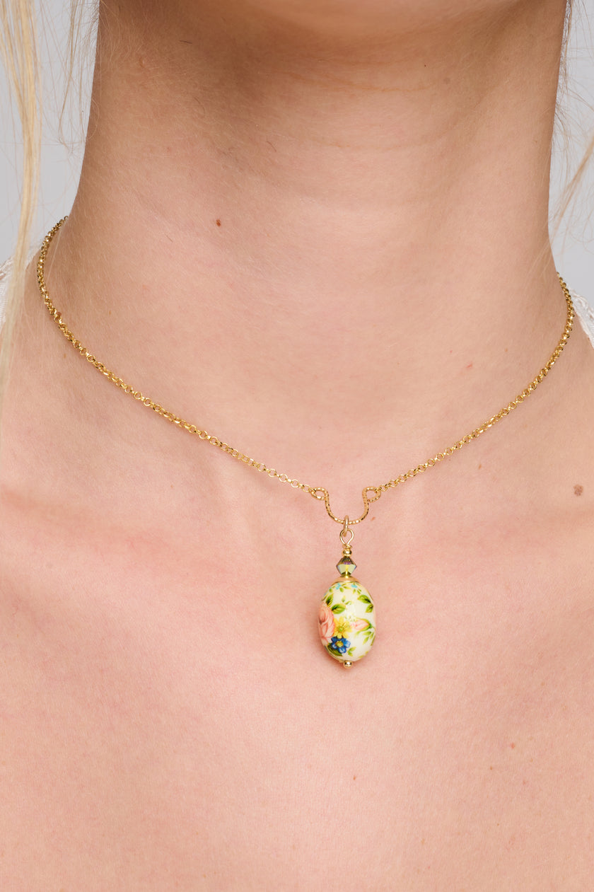 צילום של שרשרת מוזהבת עם תליון ביצה מעוצב מחרסינה או זכוכית מצוירת ביד, הכוללת ציור ורד רך בגוון אפרסק-ורוד עם פרחים ועיטורי עלים. הצווארון מעוצב עם חיבור דקורטיבי שמוסיף אלגנטיות. השרשרת מתאימה ללבוש יומיומי או למראה רומנטי.