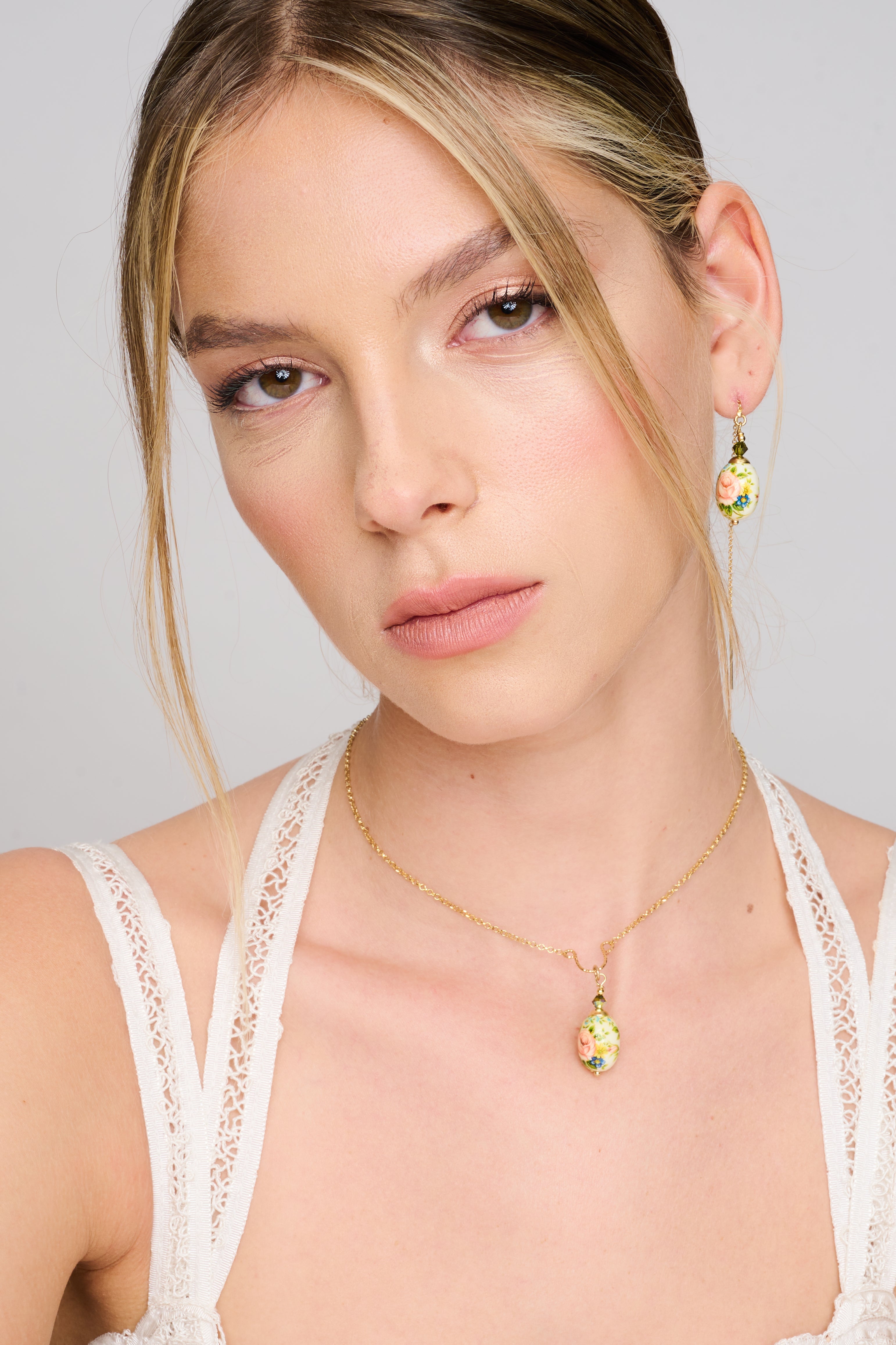 שרשרת מוזהבת עם תליון אובלי קרמי מצויר בעבודת יד, מציג ורד בגוון ורוד עדין ופרחים נוספים בכחול וירוק על רקע שנהב. החרוזים הירוקים בחיבור מוסיפים נגיעה של ברק. תכשיט בעל אופי רומנטי ונוסטלגי, המזכיר תכשיטי פורצלן עתיקים.