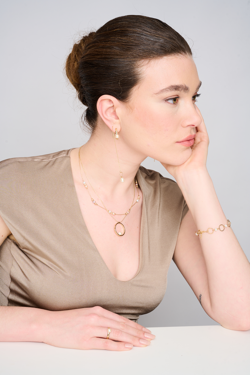 שרשרת בעיצוב אלגנטי עם אלמנט מרכזי מצופה זהב מבריק, המשובץ בפנינת שמנת מעודנת – תכשיט קלאסי בעל נוכחות בולטת. הפנינה ממוקמת במרכז התליון ומקרינה אור רך וטבעי, תוך יצירת ניגוד מחמיא עם הציפוי הזהוב. עיצוב השרשרת משלב בין עידון נשי עם נגיעה עכשווית, ומתאים לאירועים חגיגיים כמו גם ללבוש יומיומי עם נגיעה נשית. פנינים ידועות כמזוהות עם סטייל נשי טהור – ויחד עם הזהב, נוצרת הרמוניה מושלמת בין אלגנטיות לחמימות.