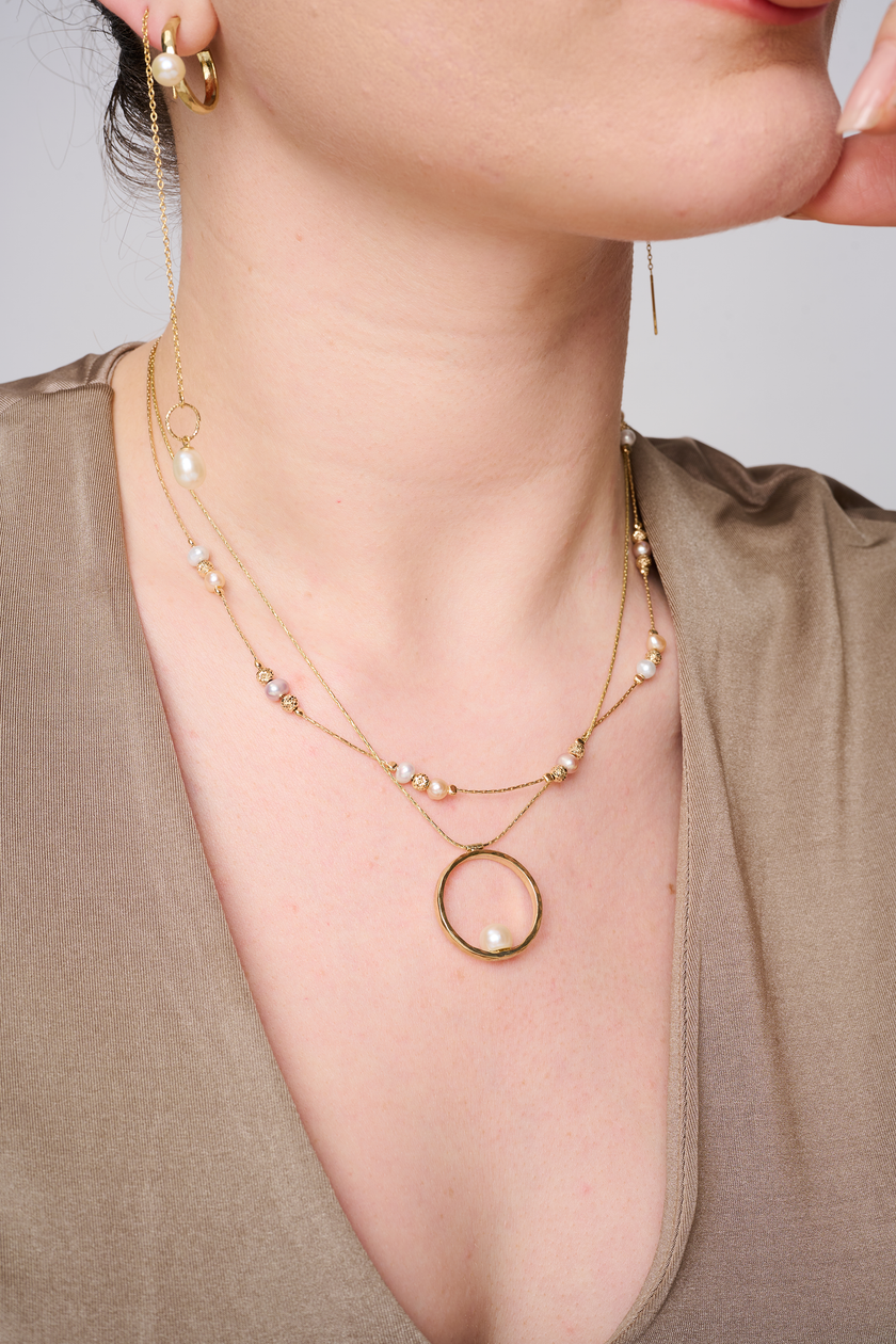 שרשרת בעיצוב אלגנטי עם אלמנט מרכזי מצופה זהב מבריק, המשובץ בפנינה לבנה קלאסית עם ברק טבעי – תכשיט קלאסי בעל נוכחות בולטת. הפנינה ממוקמת במרכז התליון ומקרינה אור רך וטבעי, תוך יצירת ניגוד מחמיא עם הציפוי הזהוב. עיצוב השרשרת משלב בין איזון בין רכות יוקרתית לאופנה עדכנית, ומתאים לאירועים חגיגיים כמו גם ללבוש יומיומי עם נגיעה נשית. פנינים ידועות כמזוהות עם סטייל נשי טהור – ויחד עם הזהב, נוצרת הרמוניה מושלמת בין אלגנטיות לחמימות.