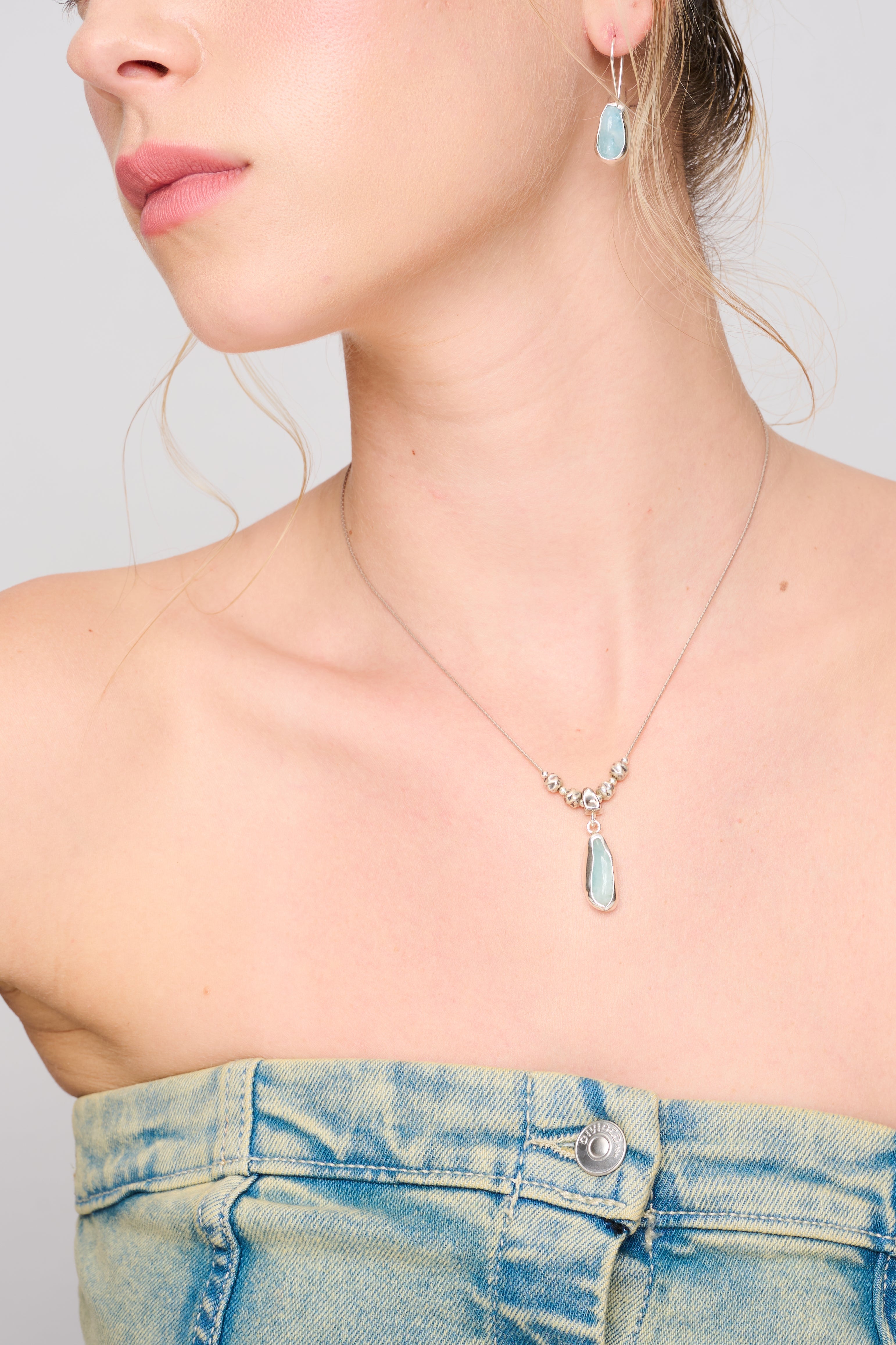 תקריב של שרשרת עדינה מכסף סטרלינג, בעיצוב מינימליסטי ומודרני. השרשרת כוללת תליון מוארך עם אבן חצי-יקרה בגוון תכלת-שקוף, מוקפת במסגרת כסף בלתי סימטרית. מעל התליון משולבים חרוזים מכסף בעבודת יד המדמים טקסטורה טבעית, להשלמת מראה אקלקטי ונקי.