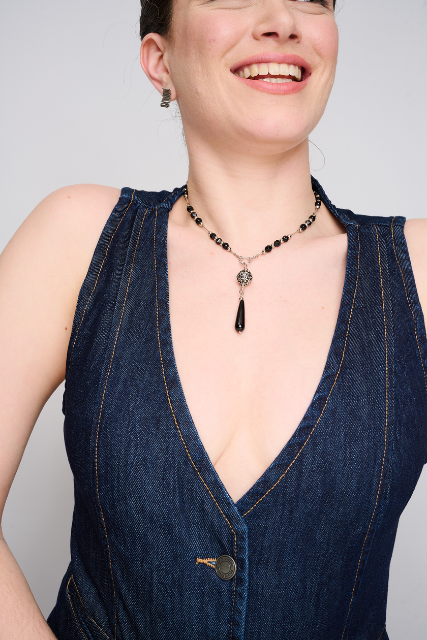 שרשרת אופנתית עם נוכחות חזקה – חרוזים עגולים באדום עז מאוניקס סינתטי, משתלבים עם חוליות כסף עדינות. תליון טיפתי מרכזי משלים את המראה הדרמטי. צילום על צוואר של אישה בלוק קליל עם חולצת ג’ינס פתוחה, המבליטה את צבע האבן.