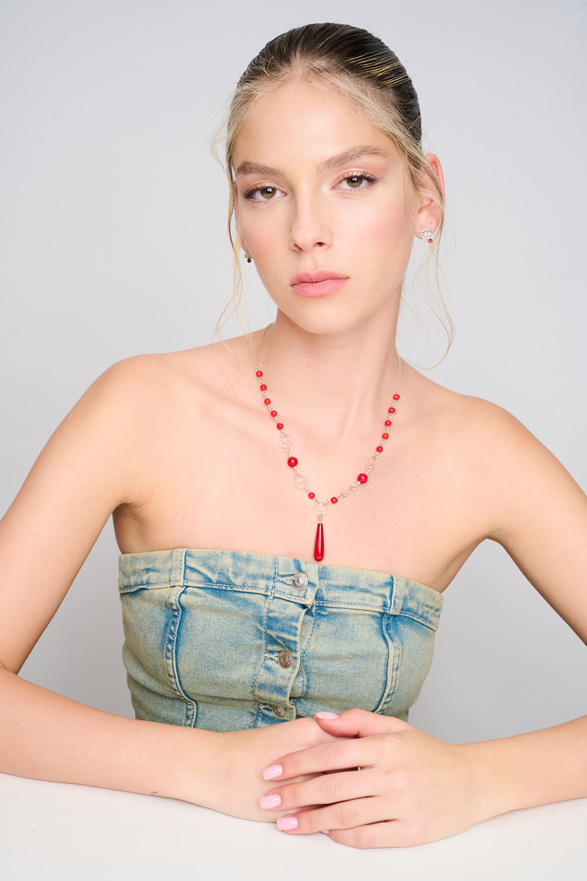 צילום תכשיט בעבודת יד: שרשרת מכסף בעיצוב משולב של חרוזים עגולים מאלמוג סינתטי אדום, טבעות דקורטיביות, ותליון טיפתי דרמטי במרכז. התמונה מדגישה את הצבע והברק של האלמוג מול גוון העור והג'ינס של הדוגמנית.