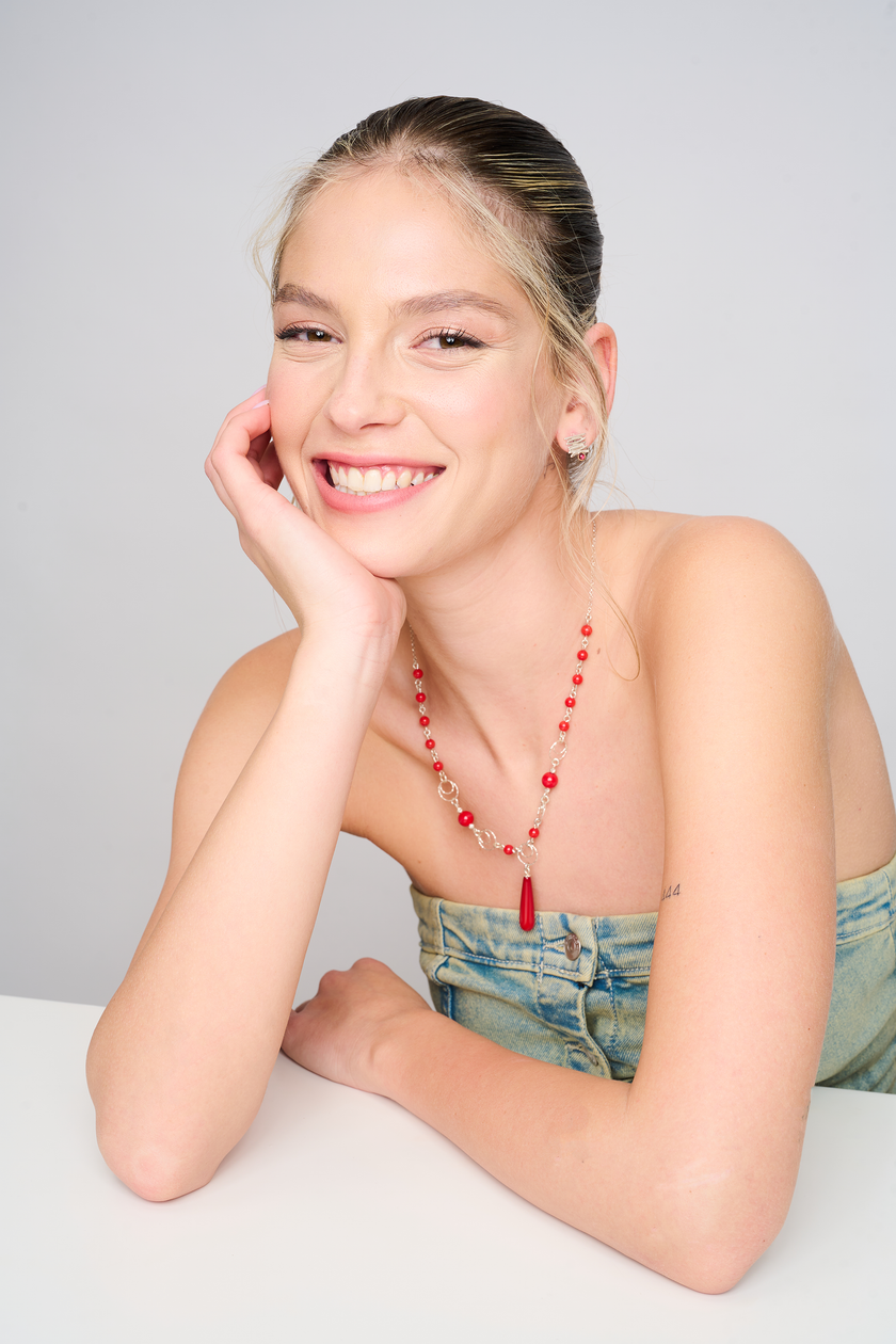 שרשרת כסף בעיצוב אלגנטי-צעיר, עם חרוזים עגולים מאלמוג סינתטי בגוון אדום חי, משובצים לסירוגין עם טבעות כסף מעודנות. במרכז תלוי תליון טיפה ארוך ומבריק. התכשיט מונח על צוואר נשי חשוף, בלבוש ג’ינס קז’ואלי, ויוצר שילוב של סטייל עם צבע עוצמתי.